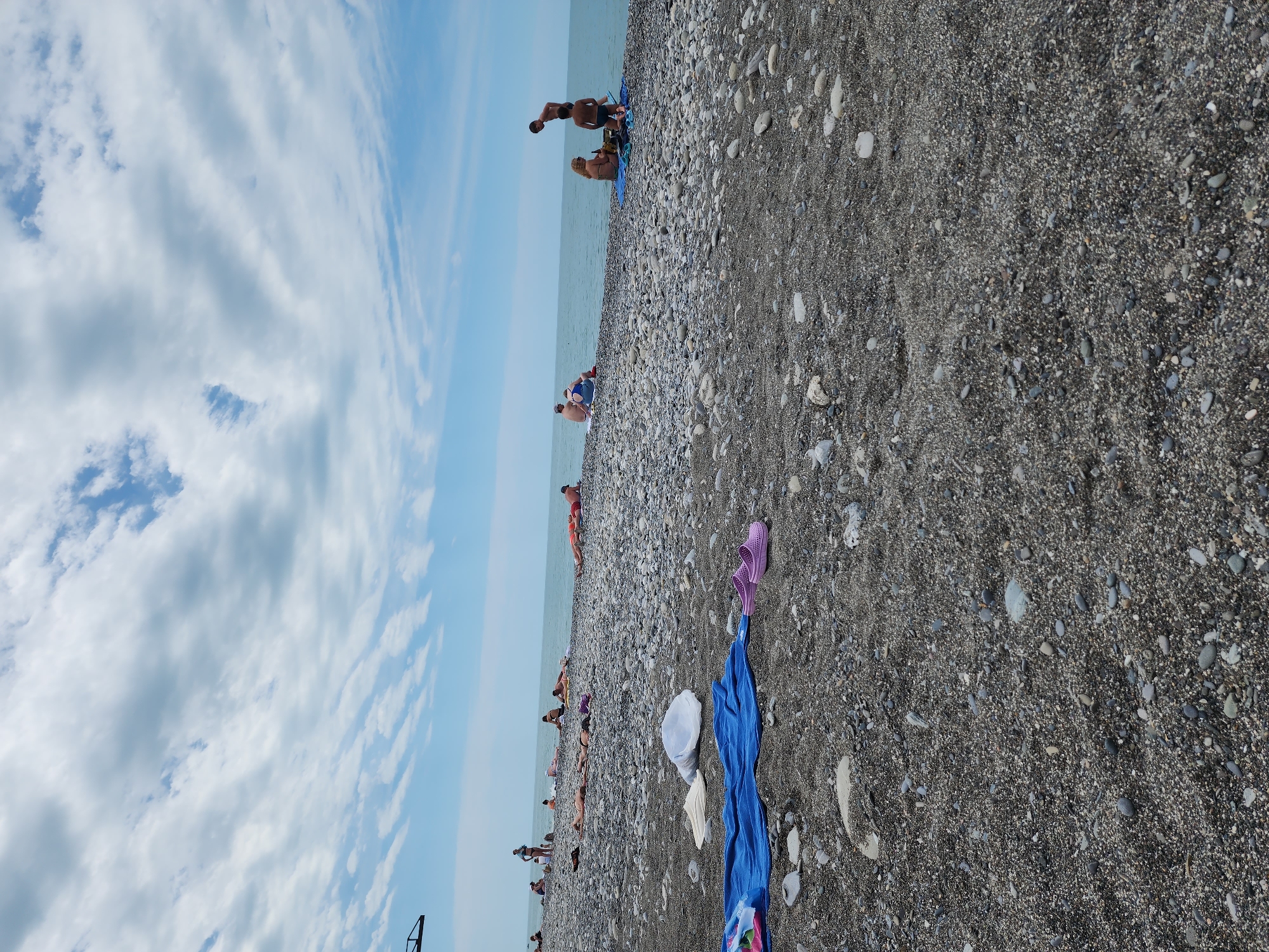 The image shows a beach on the Black Sea in Sochi, Russia. The beach is composed of pebbles and sand. People are sunbathing, swimming, and relaxing along the shoreline. Beach items such as towels and sandals are scattered on the beach. The sky is blue with scattered clouds.