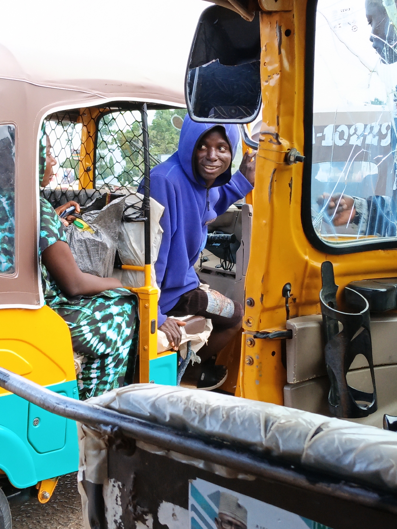 A young man wearing a blue hoodie smiles and looks towards the camera while leaning out of the driver's side of a yellow and turquoise auto-rickshaw. He is inside the vehicle, partially obscured by a black metal safety cage. To his left, a woman wearing a black, green, and blue patterned dress sits in the passenger seat. She is looking forward and to her left. The auto-rickshaw is parked on a dirt or gravel road. The scene appears to be in Bukuru, Nigeria, a town in the Jos Plateau region.