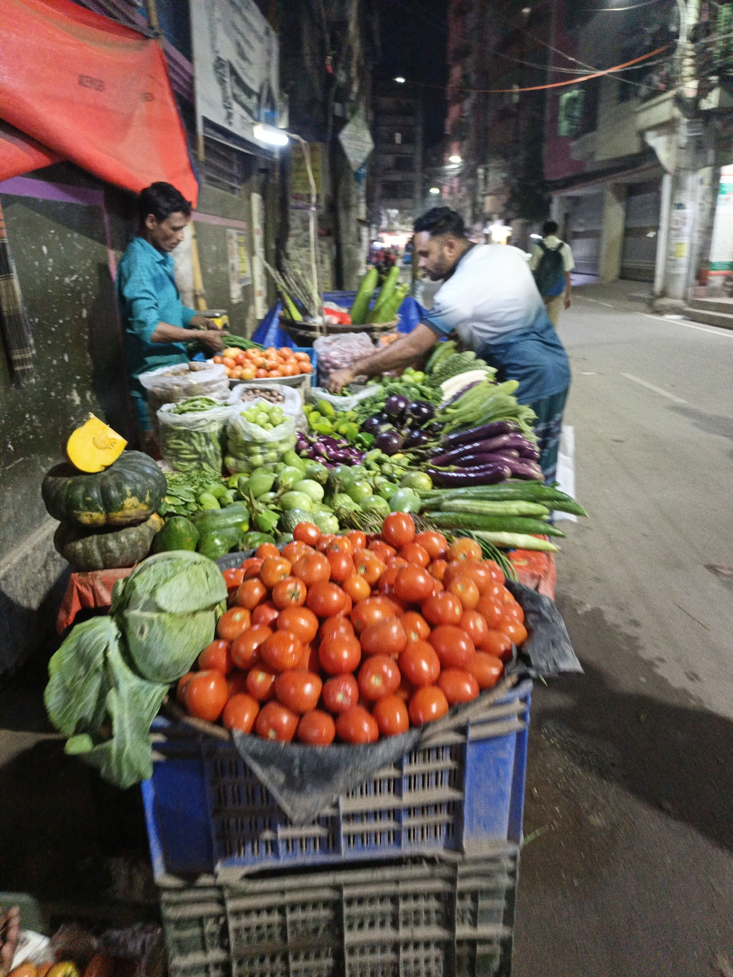 Two men are selling produce from a cart on the side of a street in Dhaka, Bangladesh. One man in a blue shirt is tending to scallions, while the other in a white and blue shirt is reaching into the produce. The cart has a variety of fruits and vegetables, including tomatoes, cucumbers, pumpkins, eggplants, cabbage, and green beans. A red tarp hangs on the left side of the image. Other people can be seen in the background, on the sidewalk. The street appears to be paved. Buildings line the street, and streetlights are visible.