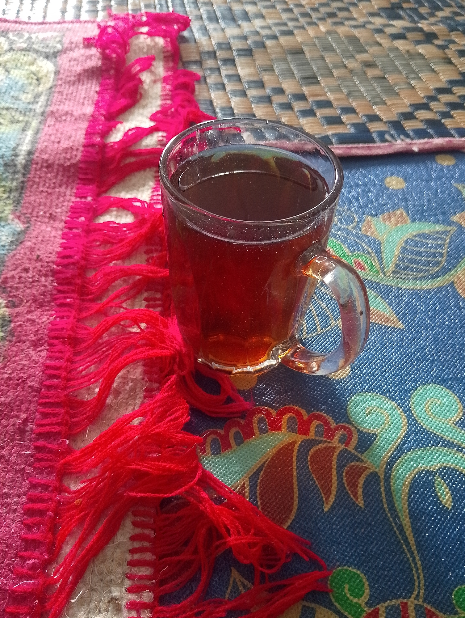 A glass mug filled with dark amber liquid, likely tea, sits on a textured blue mat with a floral pattern. To the left of the mug, a woven fabric with pink and red fringe is visible. The background consists of a woven mat with a checkered pattern of beige and blue. The image appears to be taken outdoors, possibly in a rural setting in Naga Jaya 1, Indonesia, given the natural lighting and woven textures commonly found in such environments.