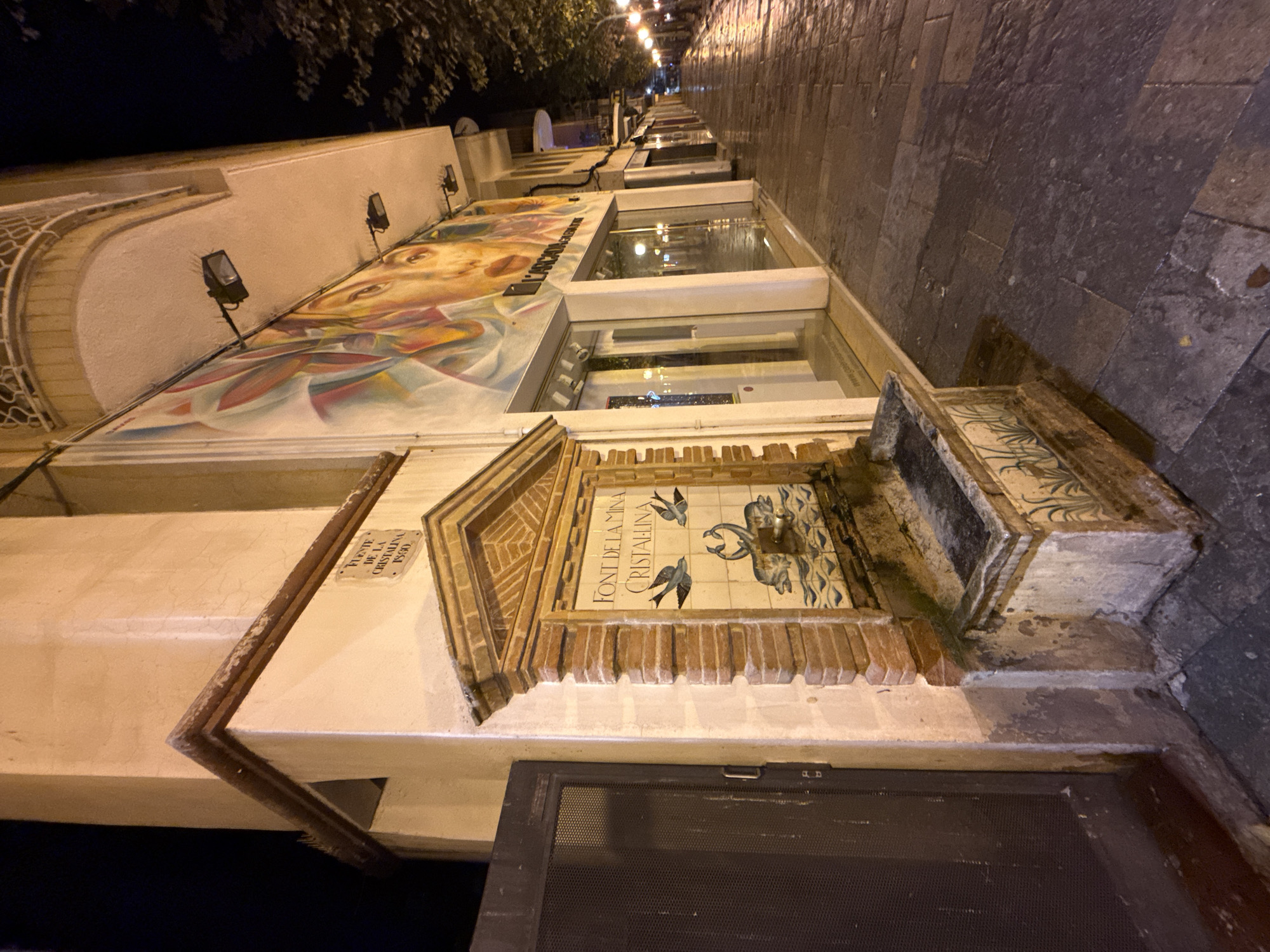 The image depicts a street scene at night in Blanes, Spain, specifically focusing on a decorative fountain attached to the side of a building. The fountain is constructed with brick and tiled panels featuring an illustration of two dolphins with their tails intertwined, creating a heart shape, beneath two birds. Above the illustration, text in Catalan reads "FONT DE LA MINA CRISTALLINA". A smaller sign above the tiled panel reads "FUENTE DE LA CRISTALINA 1880".
The building has a large mural on its facade, partially visible, depicting abstract human faces and colorful shapes. The ground level features storefronts with large glass windows, one of which displays illuminated text. The street is paved with cobblestones and appears wet, suggesting recent rain. Streetlights illuminate the scene, casting long shadows and reflecting off the wet pavement. A dark metal gate is visible to the left of the fountain. The overall atmosphere is that of a quiet, nighttime urban environment.