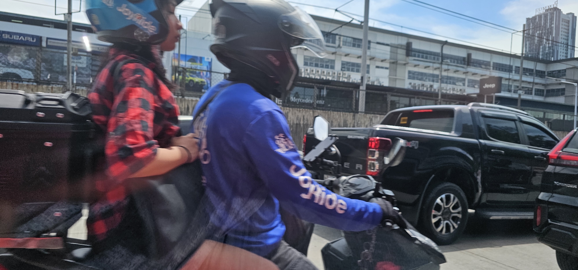 The image depicts a street scene in Quezon City, Philippines, likely on a bright, sunny day. In the foreground, two individuals are riding on a motorcycle. The person in front, wearing a blue helmet with "JoyRide" branding, is partially visible. Behind them, another person, the driver, is wearing a black helmet and a blue long-sleeved shirt with white lettering that appears to say "JoyRide" on the sleeve. They are both blurred due to motion. The motorcycle has a black storage box attached to the rear.
In the mid-ground, several vehicles are present, including a black pickup truck that appears to be a Ford Ranger, identifiable by the "4x4" badging on its rear quarter panel. Next to the pickup truck, the rear of a black SUV is visible, possibly an Xpander, with its taillight illuminated. The background reveals a commercial area with several buildings. A building with "Jeep" signage is visible, as is another taller building labeled "Summit" at the top. Further behind, a multi-story building with numerous windows and a grid-like facade can be seen, possibly a commercial or office building. The sky is clear blue with a few wispy clouds. The overall impression is a busy urban environment with ongoing traffic.