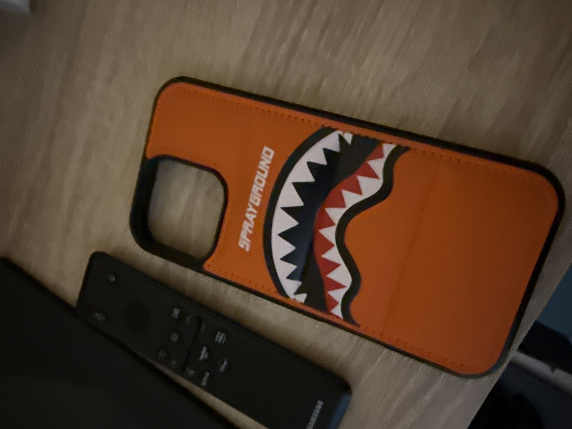 A high-angle shot shows a Samsung remote lying next to an orange Sprayground phone case on a light wooden surface. The phone case features a cartoon shark mouth design with white teeth, black gums, and a red inner mouth. The room is dimly lit. The location is not discernible from the provided image.