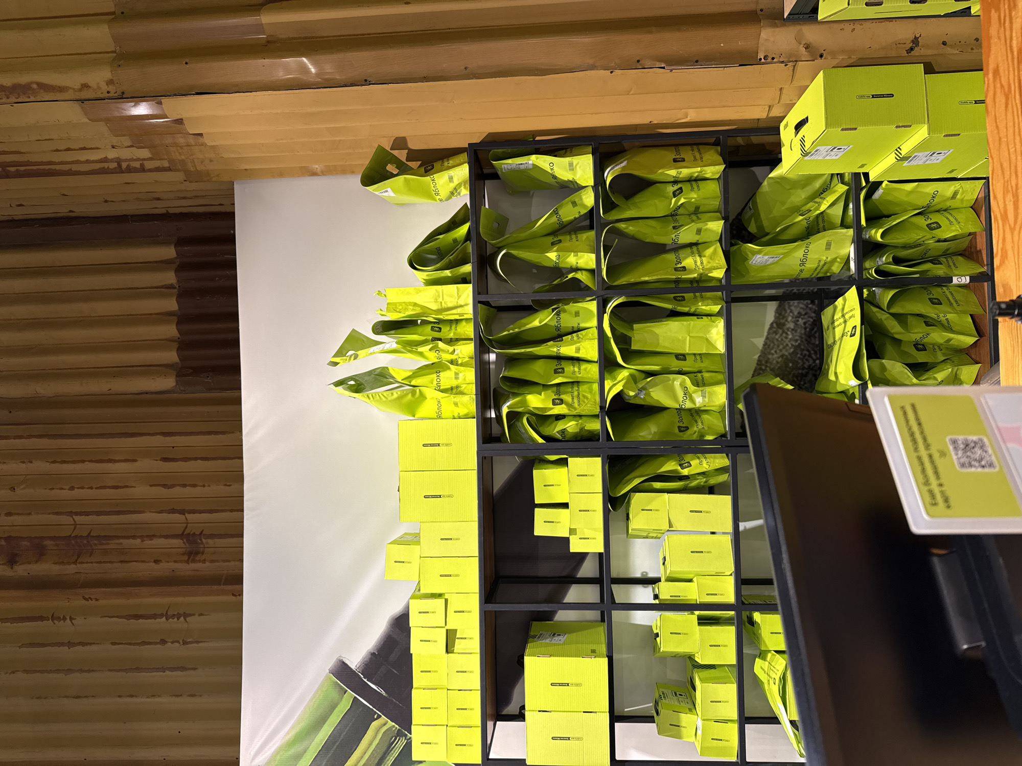 The image shows a retail display rack filled with various products, primarily in shades of yellow and green, indicative of a specific branding or product line. Bags and boxes, many labeled with text in a Cyrillic alphabet (likely Russian), fill the shelves of a black metal rack. The items appear to be merchandise for a store or brand, possibly related to food or local products. The background includes a wall, part of which is white, and the rest has a horizontal corrugated texture. A partial view of a black electronic display is in the foreground, with a pricing tag. Based on the Cyrillic text and information on the product packaging, the image is likely from Saint Petersburg, Russia.