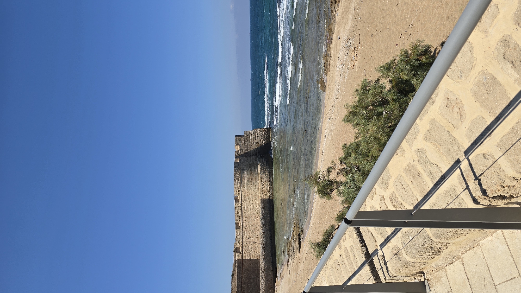 A wide shot captures the ancient stone walls of the Acre, Israel Crusader fortifications meeting the clear blue waters of the Mediterranean Sea. The walls are constructed from large, beige-colored stones and feature a curved bastion protruding into the sea. A sandy beach with scattered rocks lies at the base of the fortifications, with gentle waves lapping at the shore. In the foreground, a modern walkway made of paving stones is bordered by a gray metal railing, casting long shadows on the walkway. Green, bushy vegetation grows along the edge of the beach and near the walkway. The sky above is a vibrant, cloudless blue.