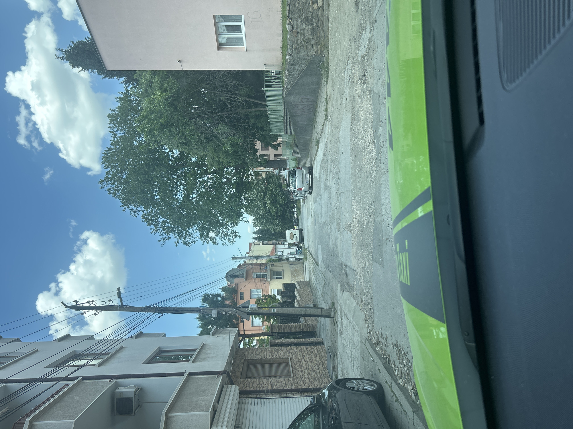 The image shows a street in a residential area of Unknown, Unknown, taken from inside a vehicle, likely a taxi given the visible "Taxi" lettering. The street is lined with buildings of varying heights and architectural styles, some appearing older with stone or brick facades, others more modern. Overhead electrical wires crisscross the sky. Several cars are parked along the street, and one white vehicle is visible further down the road. The sky is blue with scattered clouds, and a large tree dominates the center of the frame. The road surface is cracked and worn.