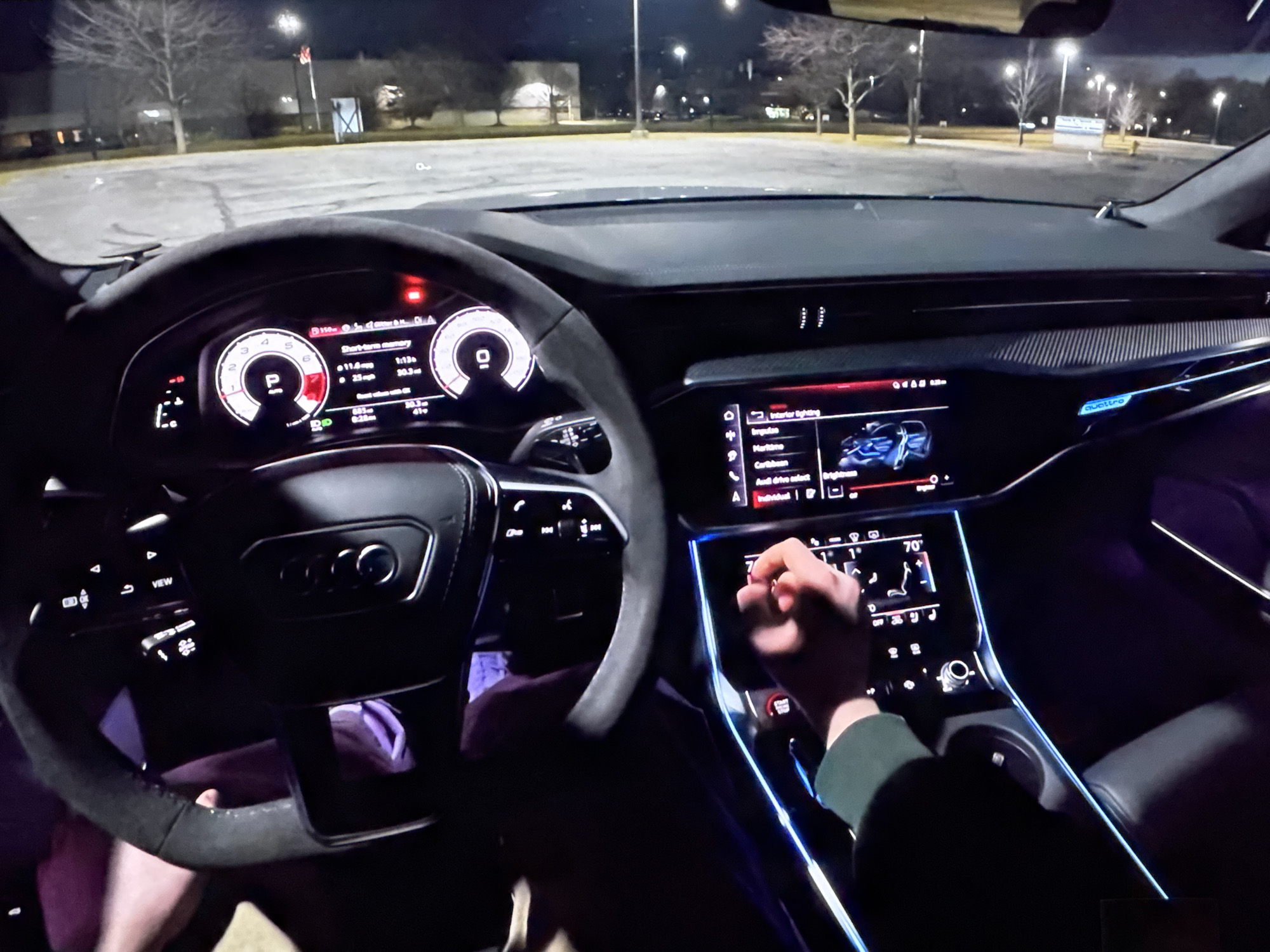 The image shows the interior of a car at night, likely in a parking lot, with the dashboard and steering wheel prominently displayed. The car's digital dashboard shows speed and other vehicle information, and the central infotainment screen is also visible, displaying interior lighting options. A person's hand is shown interacting with the touchscreen. The background reveals a dark, paved area with trees and streetlights, suggesting an urban or suburban setting. There are no identifiable landmarks that would place the image specifically in Bezden, Bulgaria. The scene is static and provides no information about ongoing events or specific activities beyond the interaction with the car's controls.