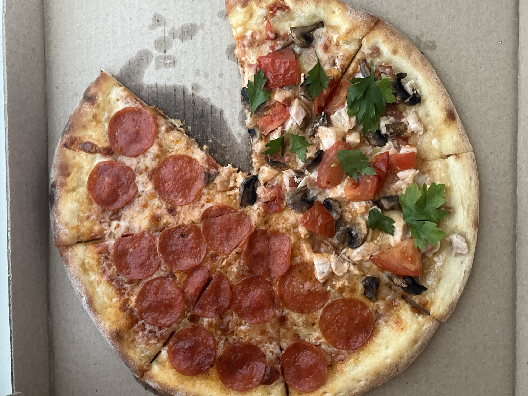 The image shows a pizza in a cardboard box. The pizza is cut into slices, and a few slices are missing. One half of the pizza is topped with pepperoni, while the other half has a variety of toppings, including mushrooms, tomatoes, chicken, and parsley. The location is not evident from the image.