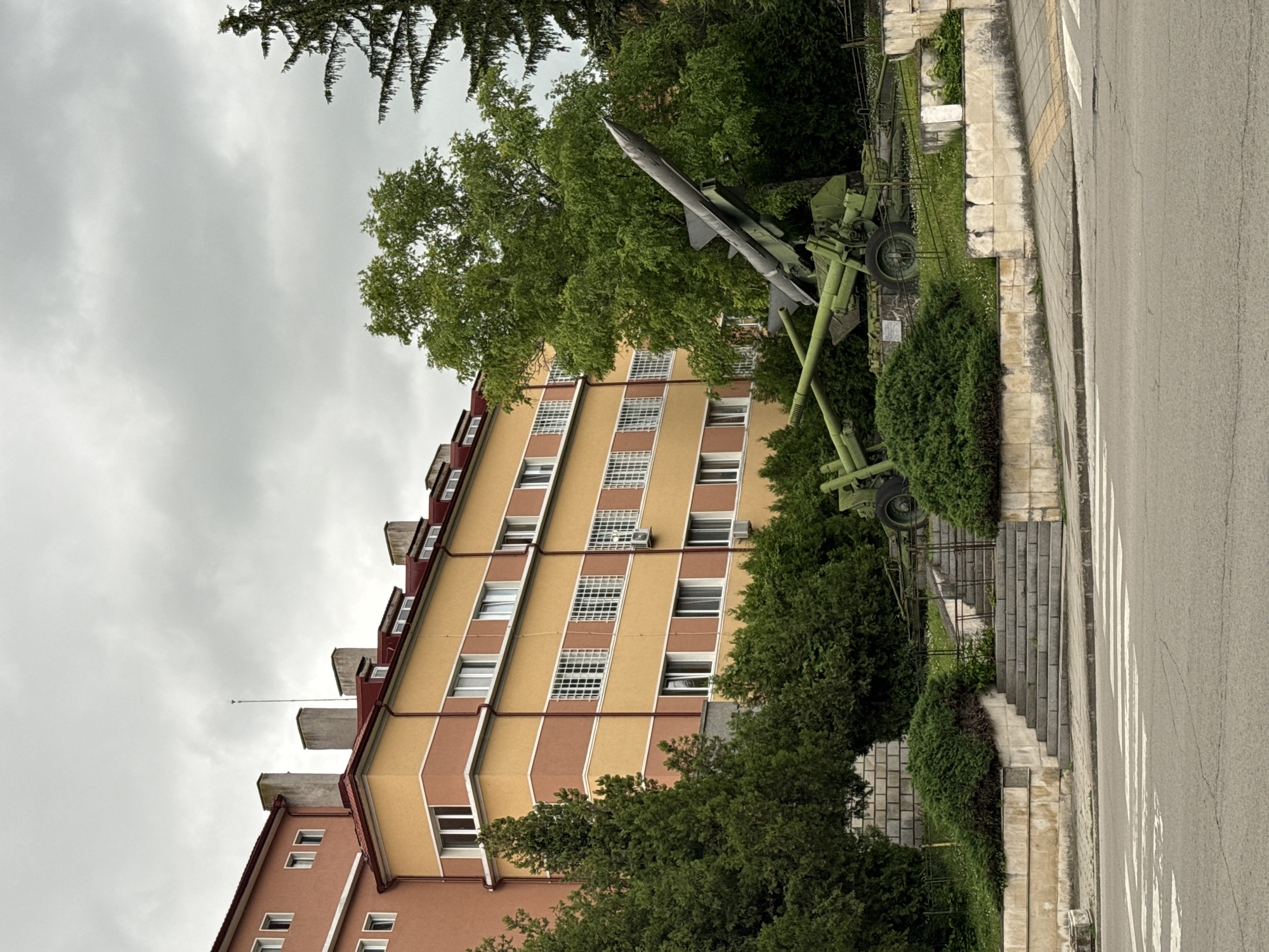 An outdoor scene in Shumen, Bulgaria, depicts a large gray missile mounted on a green military launcher as a static display or monument. The missile is angled upwards towards the left, positioned on a grassy area surrounded by green bushes and trees, which obscure parts of the launcher and ground.
Behind the monument stands a multi-story apartment building with a distinctive facade. The building features alternating vertical bands of yellow and reddish-brown, with numerous rectangular windows. Some windows, particularly those in the yellow sections, have white security grilles. The roofline of the building, visible on the left side, shows multiple dormer-like structures.
To the right of the missile display, a paved road is visible, featuring a white crosswalk. Adjacent to the road is a concrete sidewalk and a set of stone steps leading upwards into the green area where the missile is located. The sky above is overcast and gray, suggesting a cloudy day. There are no people visible, and no activity is taking place. No visible text can be discerned on any of the objects or structures.