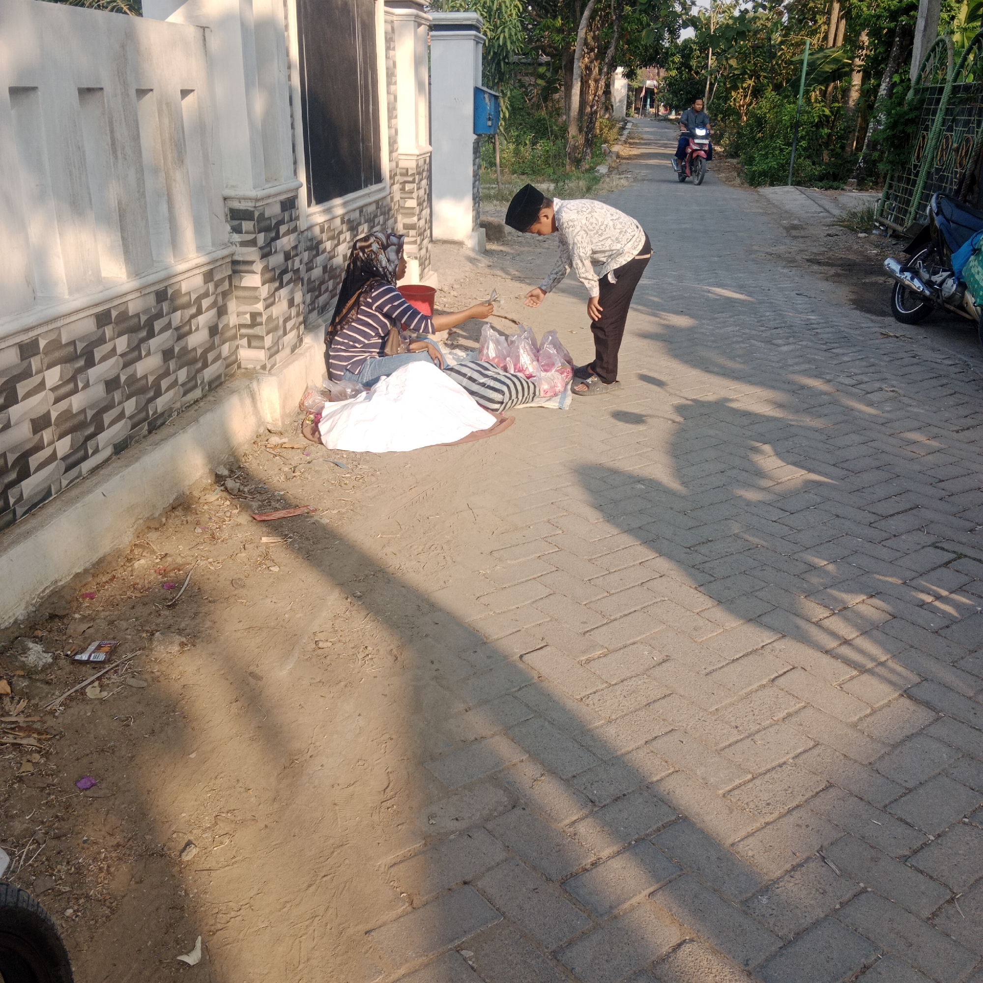In Tegowanu Kulon, Indonesia, on a paved road lined with a patterned brick wall and vegetation, a transaction is occurring. A woman, wearing a striped shirt and headscarf, sits on the ground next to a white sheet and several plastic bags filled with items, possibly snacks. She is handing something, possibly money or an item, to a young boy who is wearing a patterned shirt and a peci cap. The boy is reaching out to accept it. In the background, a man rides a motorcycle down the road. The scene is illuminated by the warm light of what appears to be late afternoon, casting long shadows across the pavement. The environment is a residential street with a mix of natural and man-made elements.
