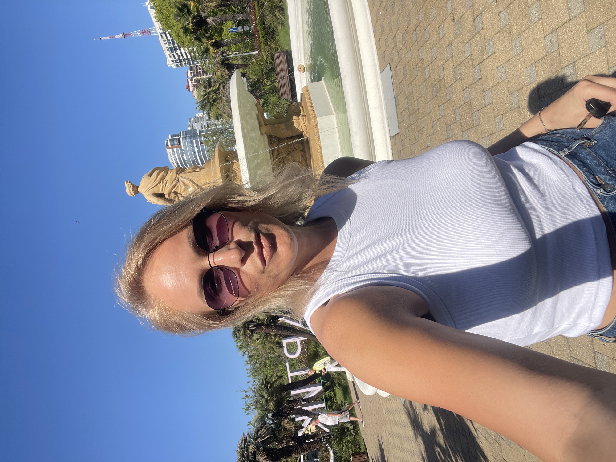 A selfie taken by a blond woman with sunglasses outside the Winter Theater in Sochi, Russia. The fountain and the theater are visible in the background, along with the Sochi cityscape and blue sky. The woman is wearing a white top and denim jeans. She is holding car keys in her hand. Palm trees are present.