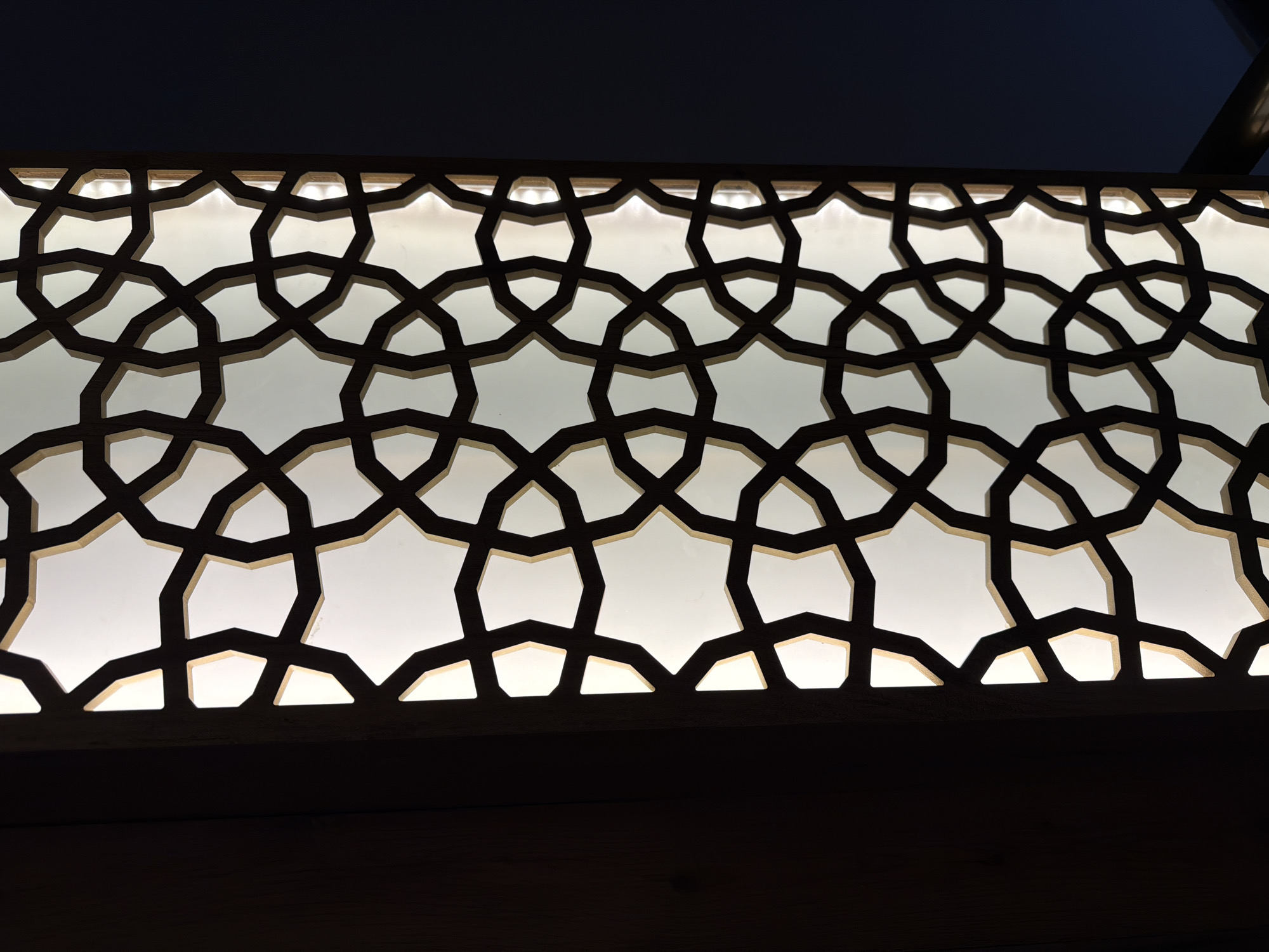 The image displays a close-up view of a decorative screen with an intricate geometric pattern. The screen is made of dark material, possibly wood or metal, and features interlocking circles and star-like shapes. Behind the screen is a translucent white panel that is illuminated from within, causing the geometric pattern to be highlighted. To the left of the screen, a dark, textured surface, likely wood, is visible. To the right, a dark, solid background, possibly a wall or curtain, creates a contrast with the illuminated screen. The lighting suggests an indoor setting, possibly a lamp or a decorative window feature. There are no people or animals present in the image, and the focus is entirely on the architectural or decorative element. The scene is static and devoid of any action.