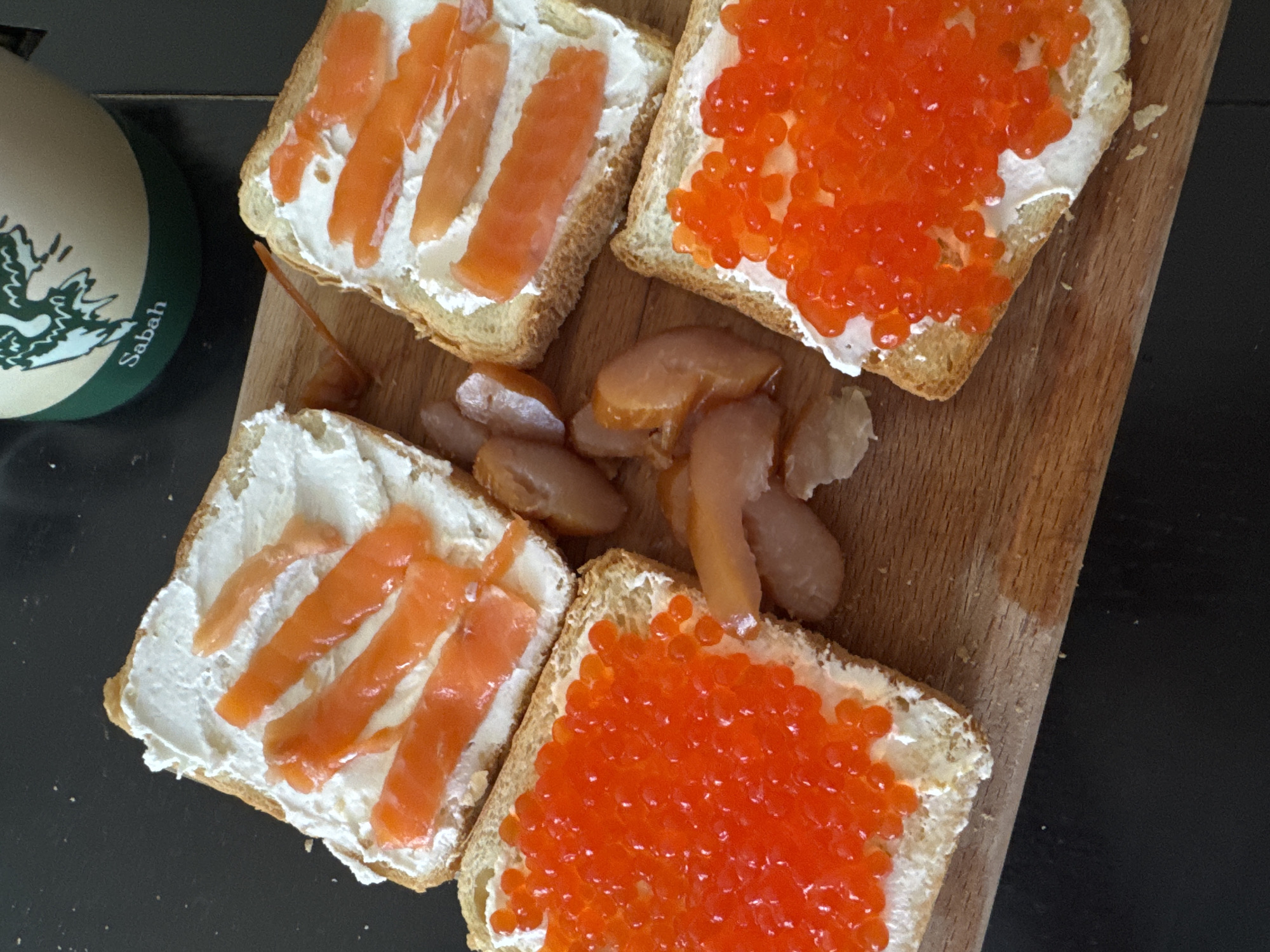 The image shows a wooden cutting board on a black surface. The board holds four square pieces of toasted bread with cream cheese spread. Two slices are topped with strips of salmon, while the other two are covered with red caviar. In the center of the board are several pieces of smoked fish. A green and white coaster with the word "Sabah" is visible in the upper left corner of the image. There is no evidence indicating this image was taken in the city of Perm, Russia.