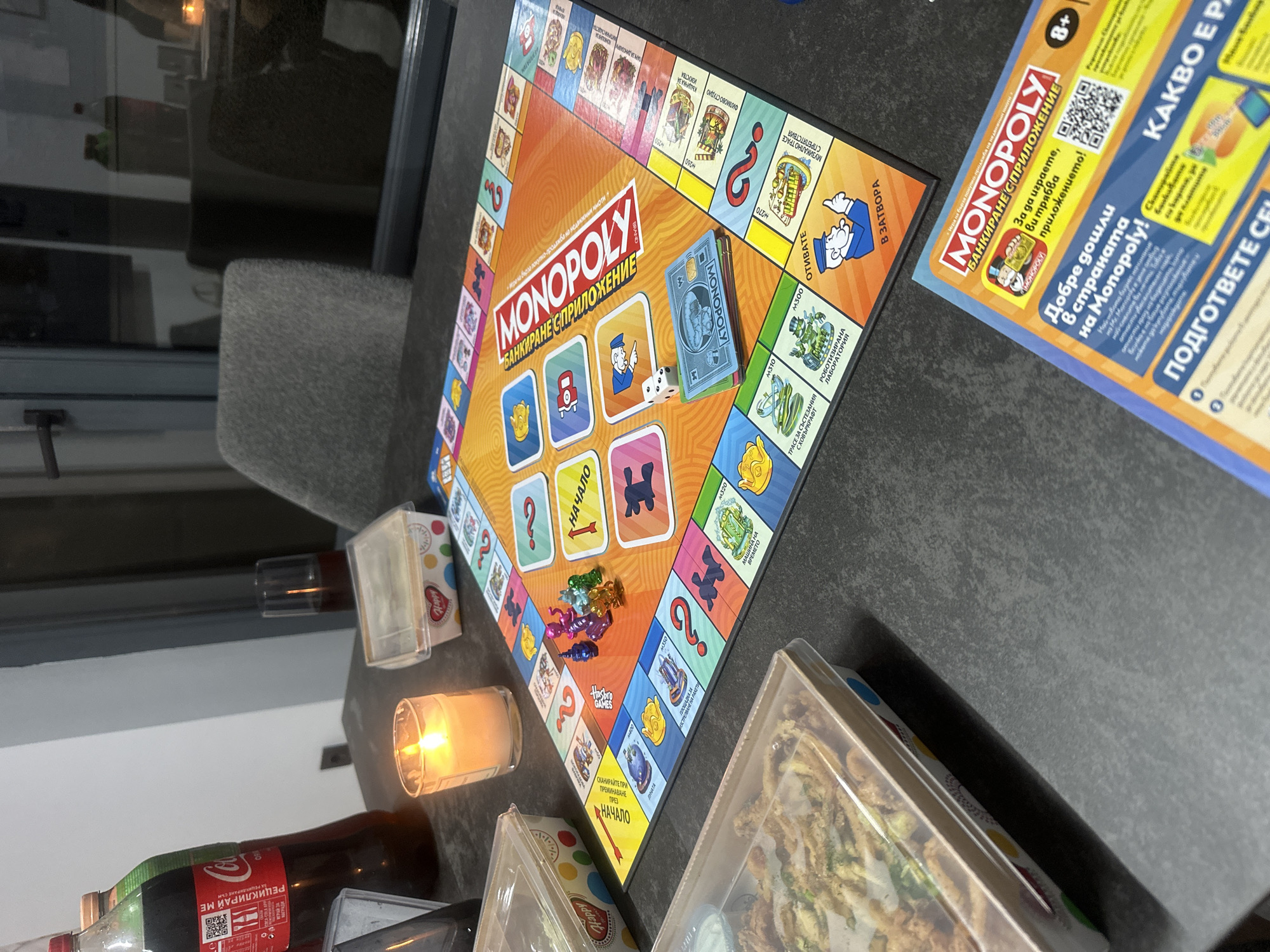 This is a close-up, eye-level shot of a Monopoly board game set up on a dark, textured surface, possibly a table. The scene appears to be indoors, with a modern, minimalist aesthetic. The background includes a large window or glass door reflecting a dimly lit interior with furniture, and a glimpse of an outdoor area with a faint light source.
Dominating the foreground is the vibrant Monopoly board, its orange and yellow hues prominent. Several game pieces, including small, colorful tokens and two dice, are scattered on the board. A stack of Monopoly money, featuring the iconic Monopoly man, is also visible. To the left of the board, a lit white candle in a glass holder casts a warm glow.
Adjacent to the Monopoly board, there are three clear plastic containers holding food, possibly takeout or a casual meal. One container in the foreground is filled with what appears to be fried food, possibly calamari or some kind of fried fish, with a creamy sauce on the side. The other two containers, further back, are less clearly visible but appear to contain similar food items.
On the right side of the image, a portion of a Monopoly board game box or promotional material is visible, featuring the Monopoly logo and text in Bulgarian. This suggests the game is a specific edition, possibly related to an application or a special theme.
There are no people directly visible in the image, but the presence of the food and the game suggests a social gathering or a relaxed evening at home. The lighting is a mix of ambient light from the background and the direct light from the candle, creating soft shadows.
The presence of Bulgarian text on the Monopoly box and game board indicates the location is likely in Bulgaria. While the specific city of Ruse is mentioned, there are no definitive visual cues in the image itself to pinpoint this exact location. However, the context of a home or dining setting with a Monopoly game is evident.