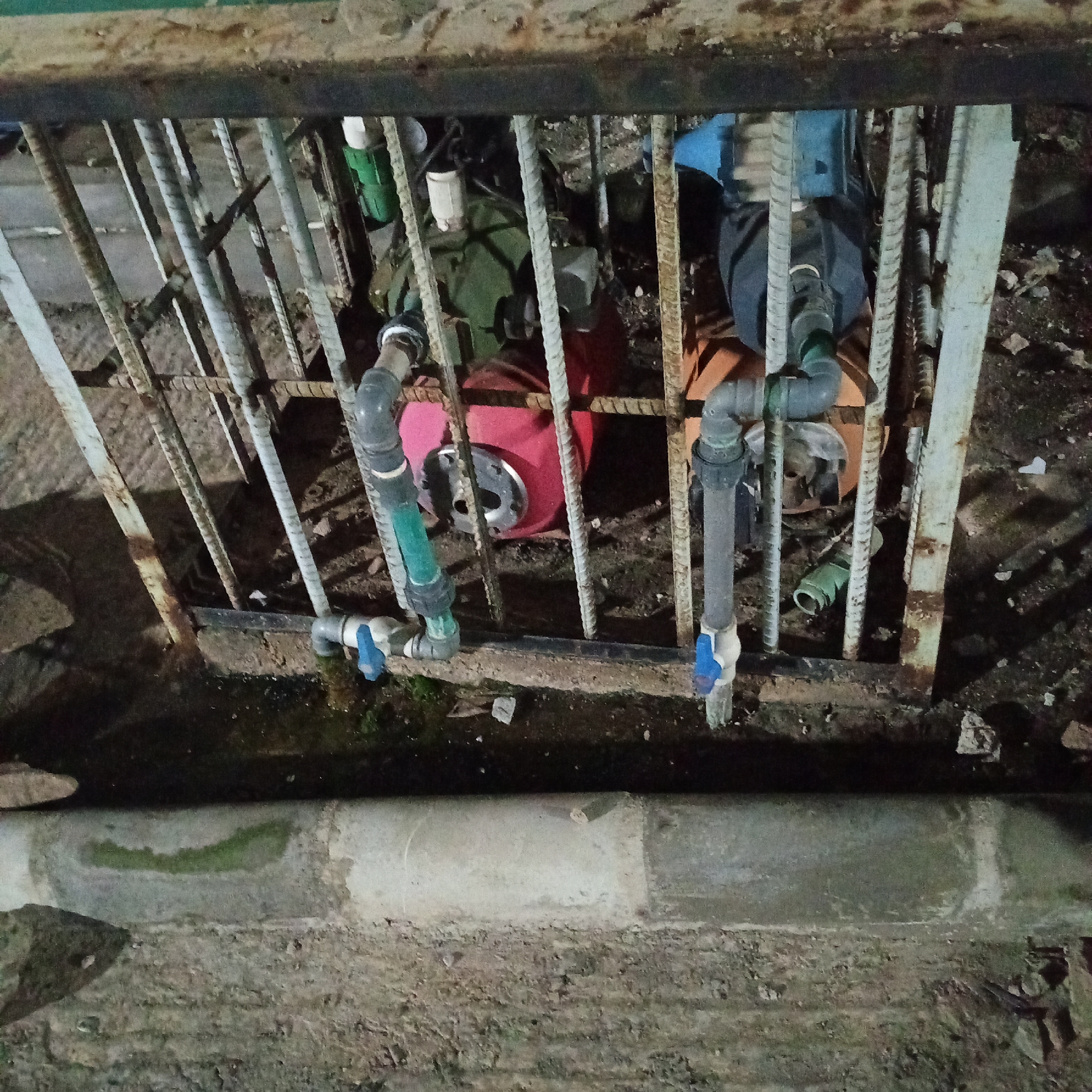 The image shows an outdoor scene with pipes and mechanical equipment, likely part of a water pump system, enclosed within a rusty metal cage. The ground is uneven and appears to be made of dirt and rubble. There are some plastic pipes and what looks like a red water filter or pump housing visible. A concrete structure runs along the bottom of the image, possibly a drainage channel or curb. The overall impression is one of disrepair and neglect.
This scene could be located in a less developed area of the city of Ibadan, Nigeria, perhaps in a residential backyard, a communal space, or an industrial area. The presence of exposed and somewhat dilapidated infrastructure like the metal cage and rusty bars suggests a functional but not well-maintained installation. The dirt and rubble on the ground, combined with the visible pipes and equipment, point towards an area where practical, rather than aesthetic, concerns dominate. This could be a location where water is sourced or distributed for local use, and the image captures the utilitarian nature of such a setup.