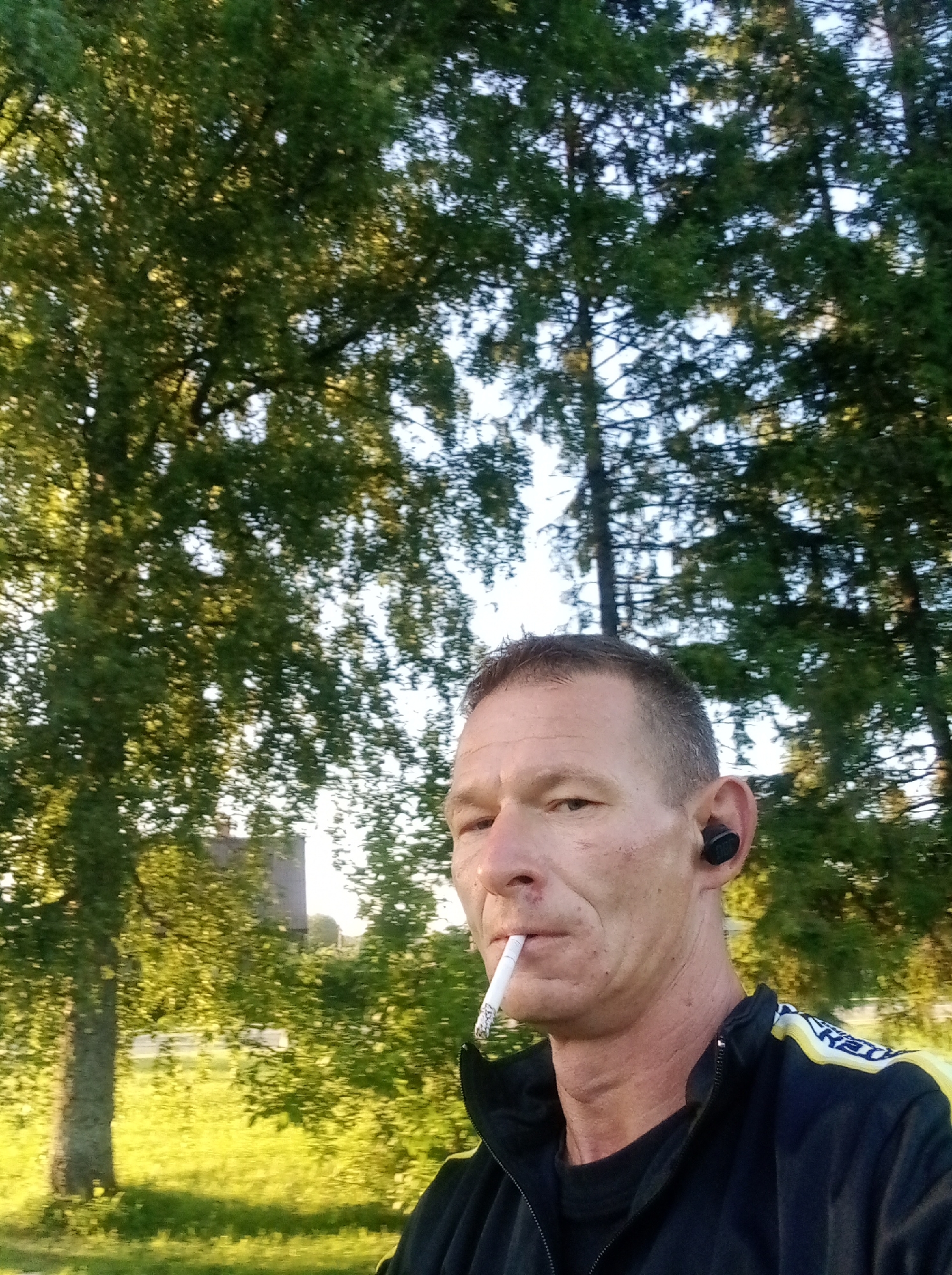A man is pictured from the chest up, smoking a cigarette, outdoors in Tapa, Estonia. He has short, grey hair and is wearing a black zipped jacket with yellow and white accents on the shoulders. A black wireless earbud is visible in his right ear. The background is filled with trees and greenery, with dappled sunlight illuminating the scene. The lighting suggests it is late afternoon or early morning.