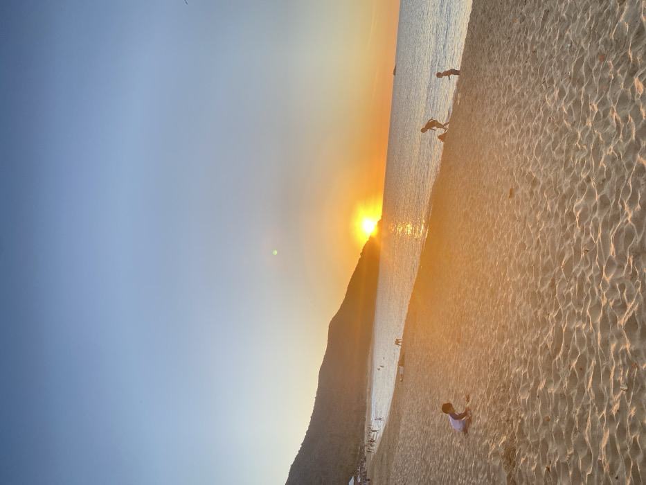 This image displays a coastal sunset scene at a sandy beach in Altagracia, Venezuela. The large, bright orange sun is setting on the horizon, partially obscured by a dark, vegetated hill on the left, casting intense golden light across the scene. A prominent lens flare is visible in the upper mid-sky. The sky transitions from light blue overhead to warm orange and yellow near the horizon. The foreground is dominated by a wide expanse of light-colored sand, showing ripples and footprints. In the lower-left foreground, a young child, appearing to be 3-6 years old, wearing a light shirt with blue sleeves and dark shorts, is kneeling and interacting with the sand. Towards the mid-ground right, two additional children are observed near the water's edge; one is bent over, and the other stands upright beside them, both silhouetted against the bright reflection on the calm sea. The water surface displays a strong golden reflection path from the setting sun. In the distant left, approximately 15-20 individuals are scattered along the shoreline and wading in the shallow water. A light-colored tent or canopy structure is visible on the far left beach, near the base of the hill. A small, indistinct vessel is present on the distant right horizon line.