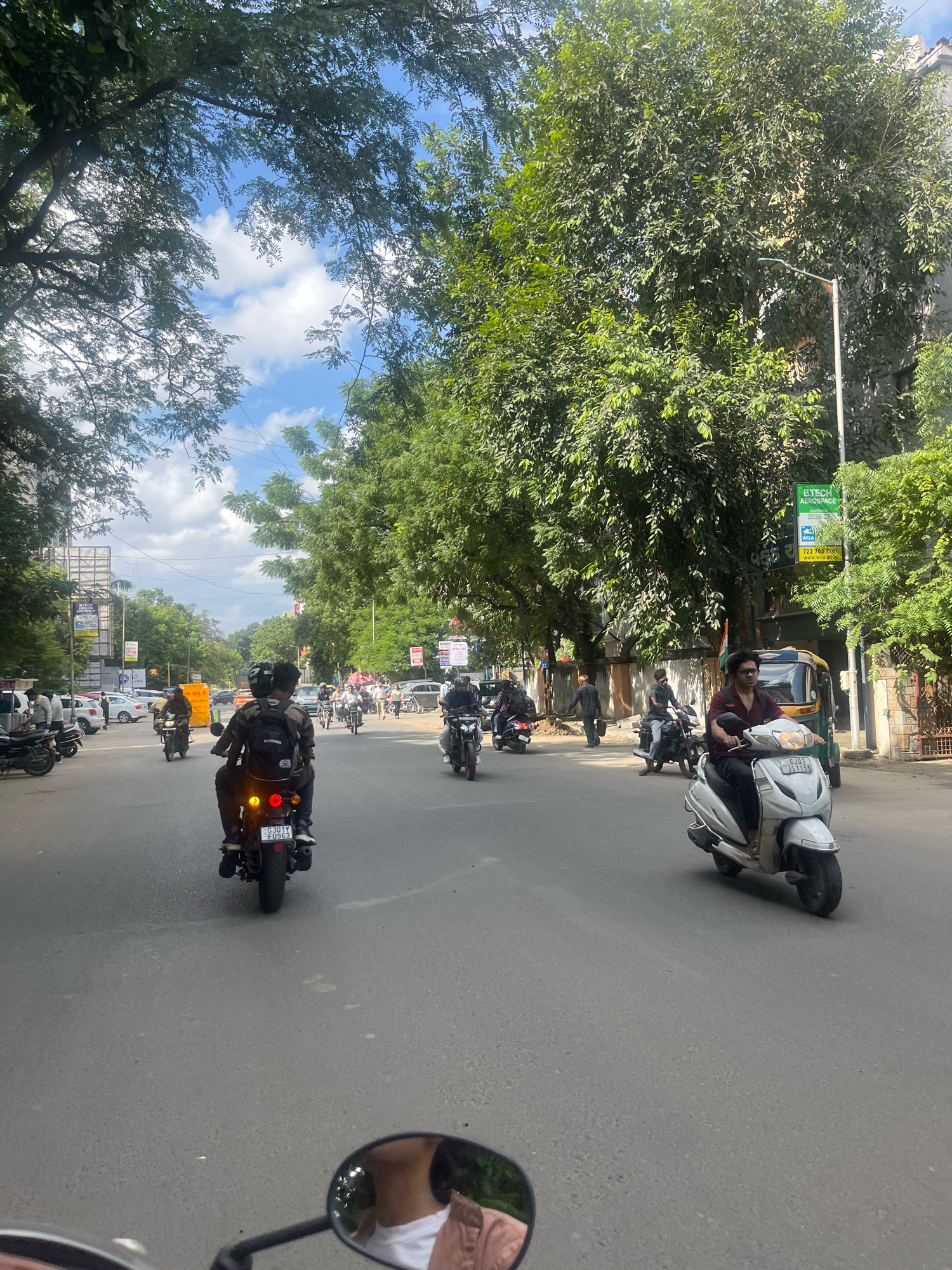 The image depicts a street scene in Navrangpura, India, with a focus on vehicular traffic and pedestrian activity under a partially cloudy sky. The perspective appears to be from a rider on a two-wheeled vehicle, with a motorcycle mirror visible in the foreground. A rider and passenger on a motorcycle are centrally positioned, heading away from the viewer, their vehicle displaying a license plate with the characters "GJ01YY F0963". To the right, a person is riding a white scooter, also moving away. Other motorcycles and a three-wheeled auto-rickshaw are also visible on the road, along with several parked cars. The street is lined with lush green trees on both sides, partially obscuring buildings. Advertising billboards and streetlights are present along the roadside. Several pedestrians are seen walking on the sidewalks and crossing the street. The overall atmosphere suggests a typical day in a bustling urban environment.