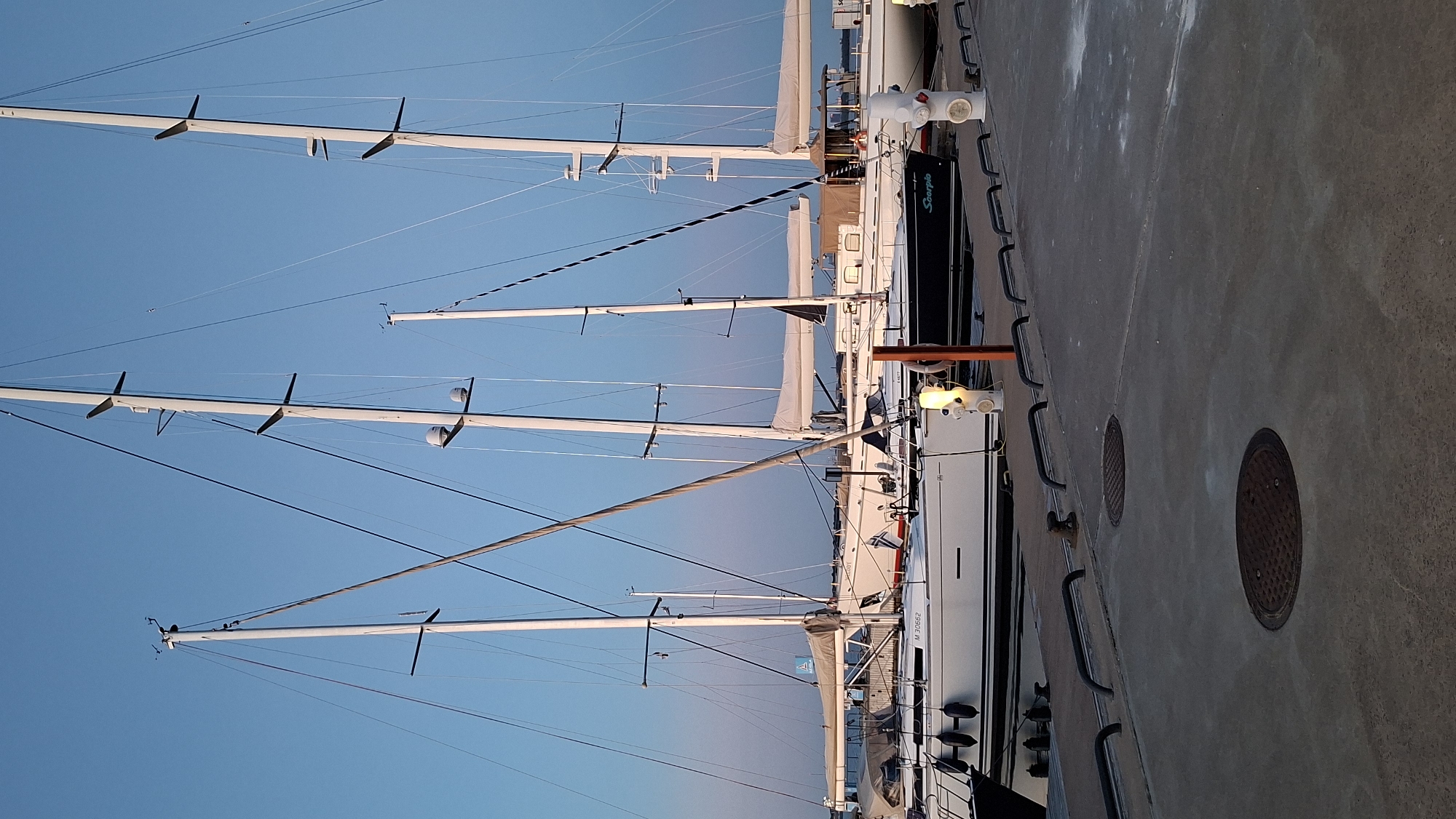 In Tallinn, Estonia, a fleet of sailboats is docked at a harbor. The boats are lined up along a concrete pier, with their masts extending towards a clear blue sky. The scene is peaceful, with the boats appearing to be at rest. Some of the boats are white, while one is a dark color with the name "Scorpio" visible on its side. The pier is equipped with bollards and railings, and two manhole covers are visible on the concrete surface. The lighting suggests it is either early morning or late evening, as the sun is low on the horizon.