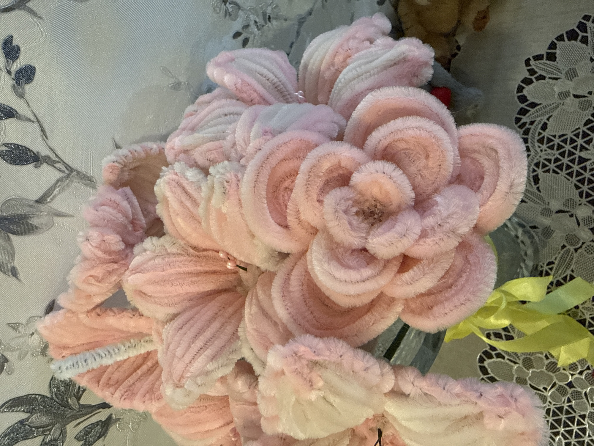 The image shows a bouquet of pink and white pipe cleaner flowers. The flowers are tightly packed, with a large central flower dominating the arrangement. A yellow ribbon is visible among the flowers on the right side. The background features a wall with a light-colored floral pattern on the left and a lacy, patterned surface on the right. No people are visible in the image. There is no evidence in the picture itself to definitively place the location as Марковский, Russia; this information seems extraneous.