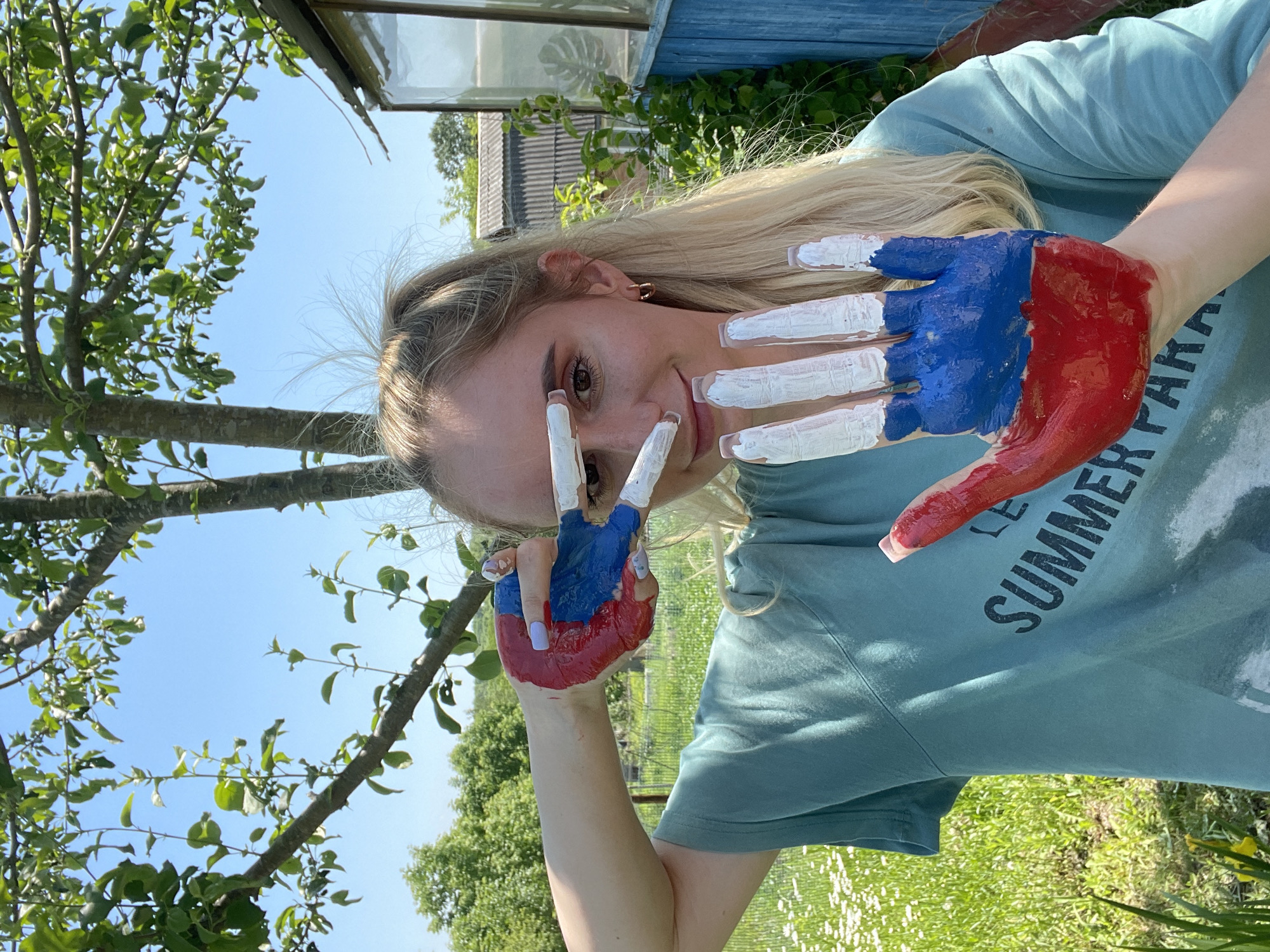 A young blonde woman stands outdoors in a grassy area with a tree overhead and a building in the background, likely Yasnoe, Russia. She is wearing a teal t-shirt with dark lettering that reads "LE SUMMER PARADISE." Her right hand is painted with the colors of the Russian flag: red on the palm and lower fingers, and blue on the upper fingers. Her left hand is also painted with red and blue on the palm and lower fingers, but the upper fingers are painted white. She is holding her hands up to her face, with the fingers of her left hand extended and partially obscuring her eyes and nose. Her right hand is held closer to her chest. The background includes green grass, white wildflowers, trees, and a blue wooden structure with glass panels. The sky is clear and blue.
