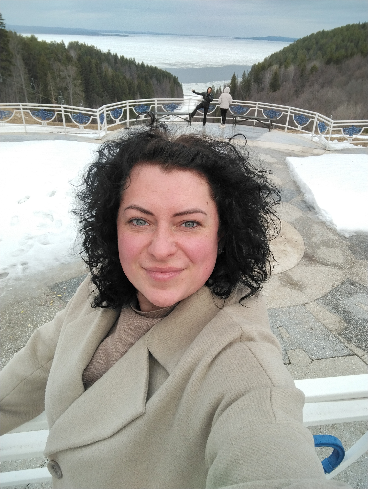 The image shows a selfie of a woman with curly black hair and blue eyes, wearing a beige coat. In the background, there is a scenic view of a lake partially covered with ice, flanked by forested hills. Two people are visible further back, standing on a viewing platform with a white railing. The ground is covered with snow. The scene is in Galevo, Russia, evidenced by the recognizable viewpoint. The woman is standing on a stone-paved area, possibly a part of the viewing platform. One of the people in the background is standing on one leg, in the air.