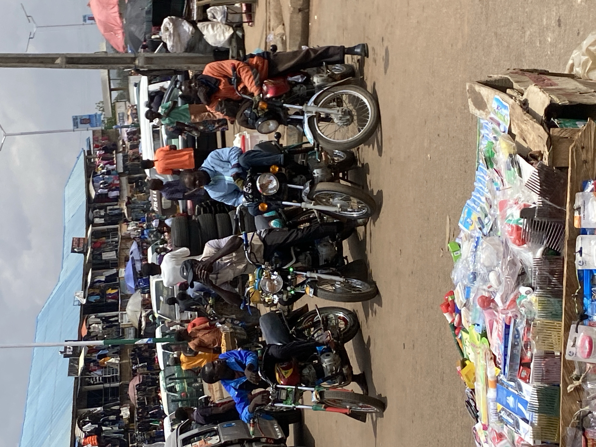 The image captures a vibrant and busy street scene, likely a market area, in Bukuru, Nigeria. The photograph, taken during daytime under clear skies, showcases a slice of daily life filled with commerce and activity.
In the foreground and midground, several men are prominently featured, most of whom are either seated on or standing next to motorcycles. These motorcycles appear to be commonly used commuter bikes, often referred to as "Okada" in the region, suggesting their role in local transportation or as a means of livelihood for the riders. The men are dressed in casual attire, including short-sleeved shirts and trousers.
To the immediate right, a makeshift market stall, constructed from stacked cardboard boxes, displays a variety of small consumer goods. These items, many individually packaged in clear plastic, include numerous combs, plastic trinkets, and other general merchandise, likely catering to everyday needs. Faintly visible text on some packaging includes "LEF PRIZE."
The background on the left reveals a bustling market street lined with multi-story buildings, some featuring blue and grey roofs. These structures house various stalls and shops, where more goods, such as clothes and stacked tires, are displayed for sale. Several market umbrellas are visible, providing shade for vendors and their wares. Numerous other people can be seen navigating the crowded street, engaging in shopping or moving between different areas. Vans or small buses are also visible, indicating public transport in the vicinity.
Notable details include utility poles and overhead wires that crisscross the sky, and a distant blue banner with "2023" printed on it, possibly related to a political event or calendar year. The street surface appears to be a mix of asphalt and dirt, common in busy market areas. The overall impression is one of a lively urban environment, characterized by the constant movement of people and goods, and the presence of informal commerce.