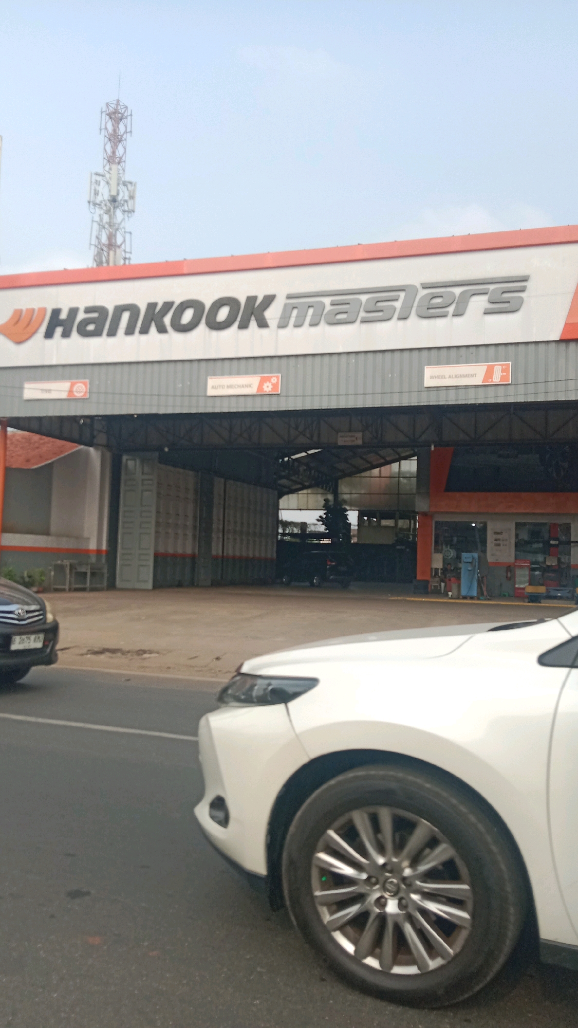 The image depicts the exterior of a Hankook Masters automotive service center located in the city of Bekasi, Indonesia. The building features a prominent white facade with the "HANKOOK masters" logo prominently displayed in black lettering. Below the logo, the building has a large, open bay area, suggesting it serves as a garage or workshop. Signs indicating "AUTO MECHANIC" and "WHEEL ALIGNMENT" are visible above the bay entrance, detailing the services offered. A telecommunications tower is visible in the distance against a hazy sky. In the foreground, the front portion of a white SUV is visible, parked on the road in front of the service center. A black car is also partially visible to the left. The environment suggests an urban or semi-urban setting, common in Bekasi.