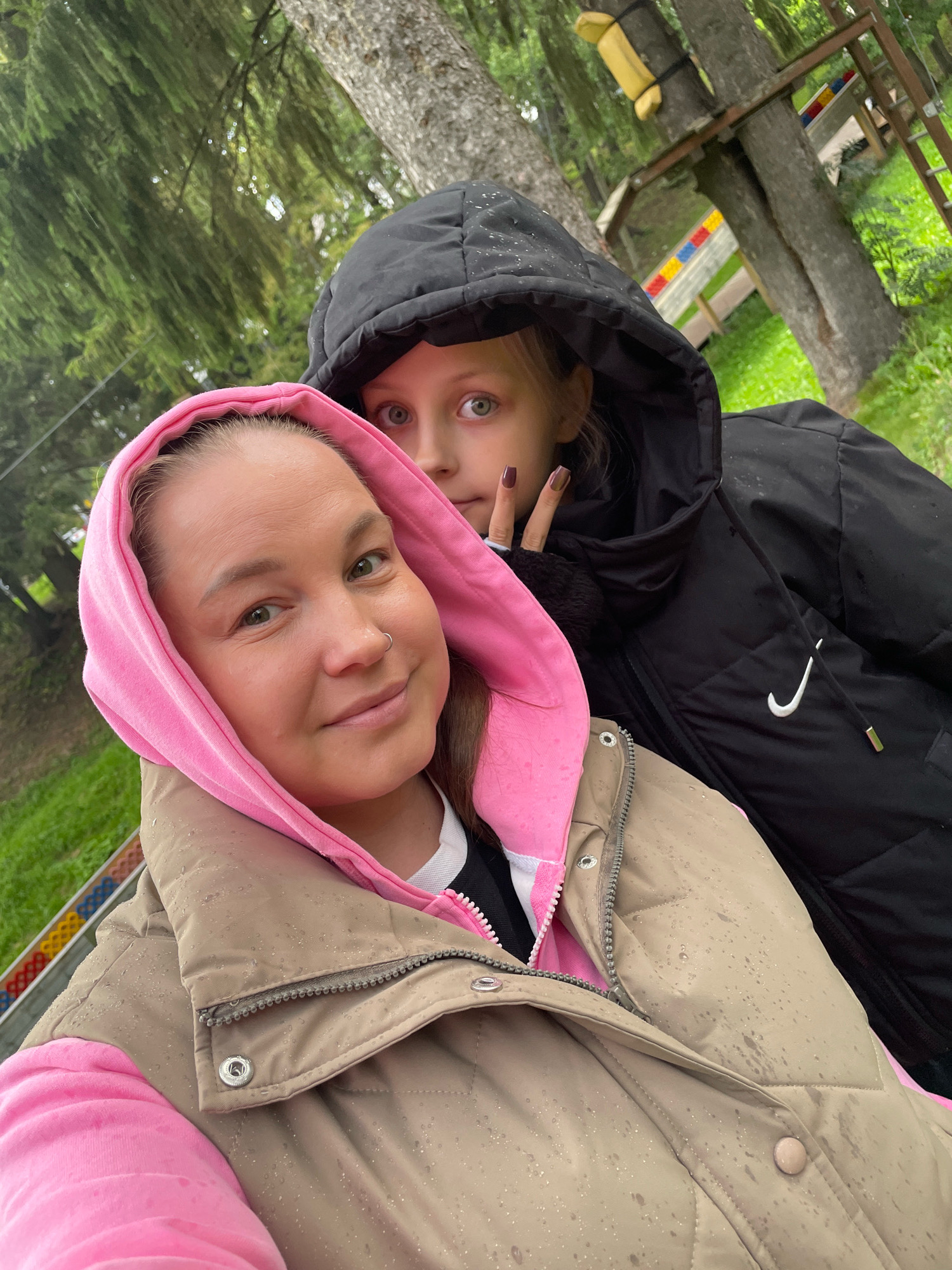 In a selfie taken outdoors, two individuals are visible. The person in the foreground, a woman, is wearing a pink hooded sweatshirt under a tan puffer vest, the vest having a textured, possibly wet, surface. She has shoulder-length brown hair and a silver hoop nose ring. To her right and slightly behind her, a child with blonde hair and wide green eyes wears a black puffer jacket with a hood. The child is making a peace sign with their right hand, which is visible near the woman's face. The background consists of trees, green grass, and what appears to be an outdoor adventure park structure with wooden beams and colorful barriers. The lighting suggests it is daytime, possibly overcast.