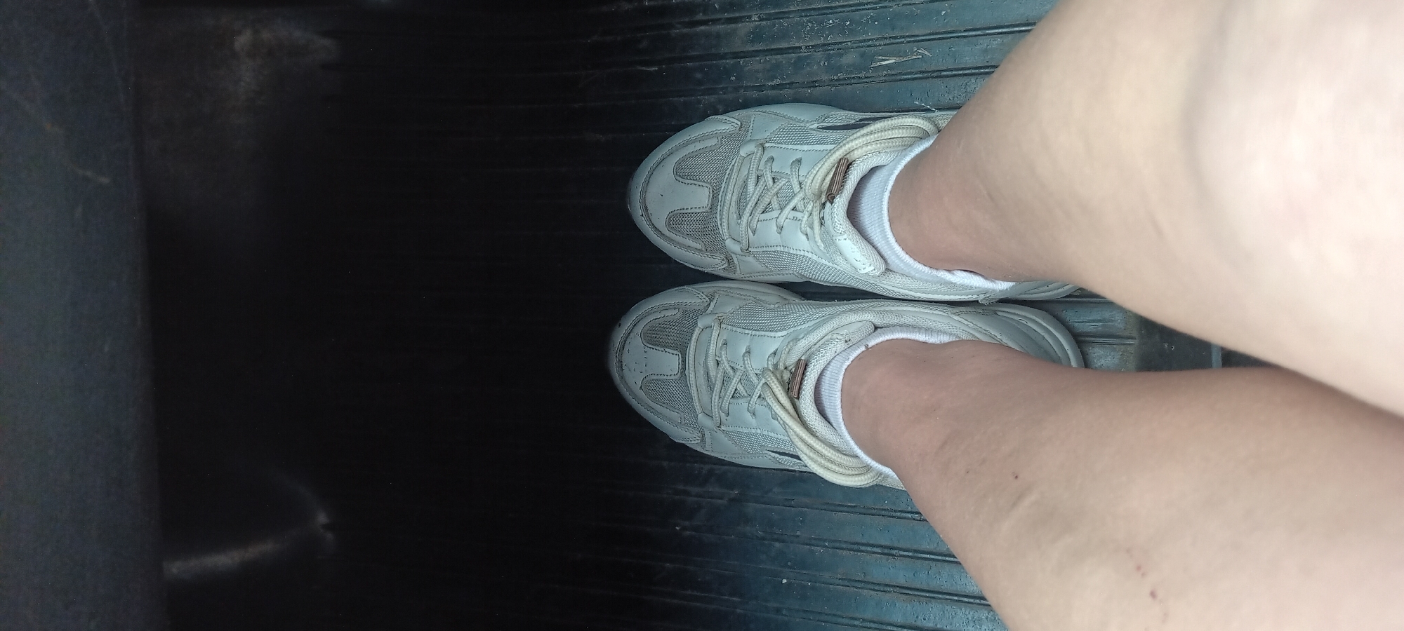 A first-person perspective shot shows a pair of lightly worn white athletic shoes with white laces against a dark, ribbed floor. The shoes are tied and the wearer is wearing white ankle socks. The legs are bare and pale, with some faint markings. The background is a dark, out-of-focus space. The image does not provide any information about the location in Koviahy, Ukraine.