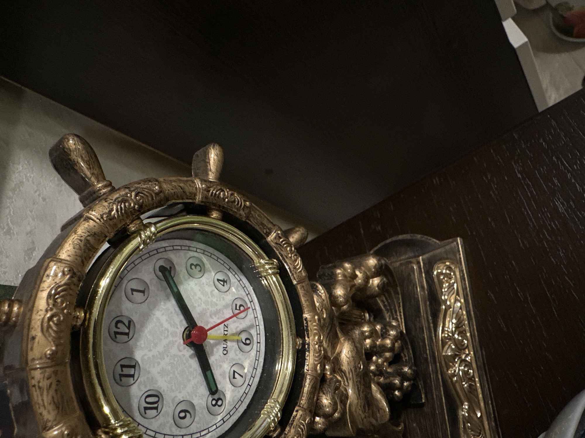 A close-up shot shows a decorative clock with a nautical theme. The clock has a ship's wheel design around the face, which is gold-colored and ornate. The clock face displays a time of approximately 11:41. The background includes parts of a dark shelf and a brown wooden surface. The location is Chaikovsky, Russia.