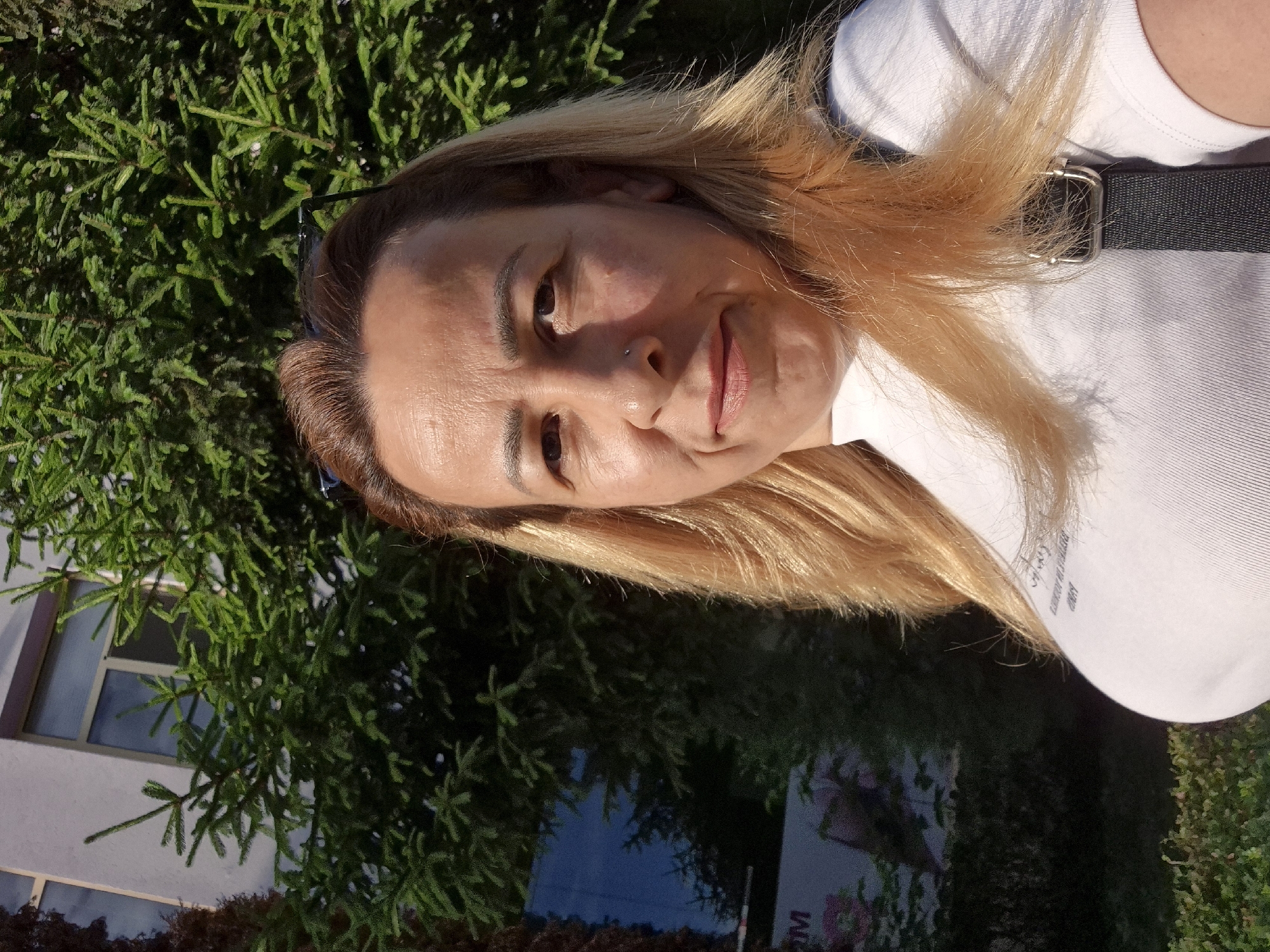 A woman with blonde hair and light-colored skin is standing outside in front of a building and some evergreen trees. She is wearing a white t-shirt with a dark strap over her shoulder. The woman is looking at the camera and smiling. The lighting suggests it is daytime, and the sun is casting shadows. There is a building with windows to her left, and a large evergreen tree behind her. Part of a sign with the letter "M" on it is visible on the left side of the image.