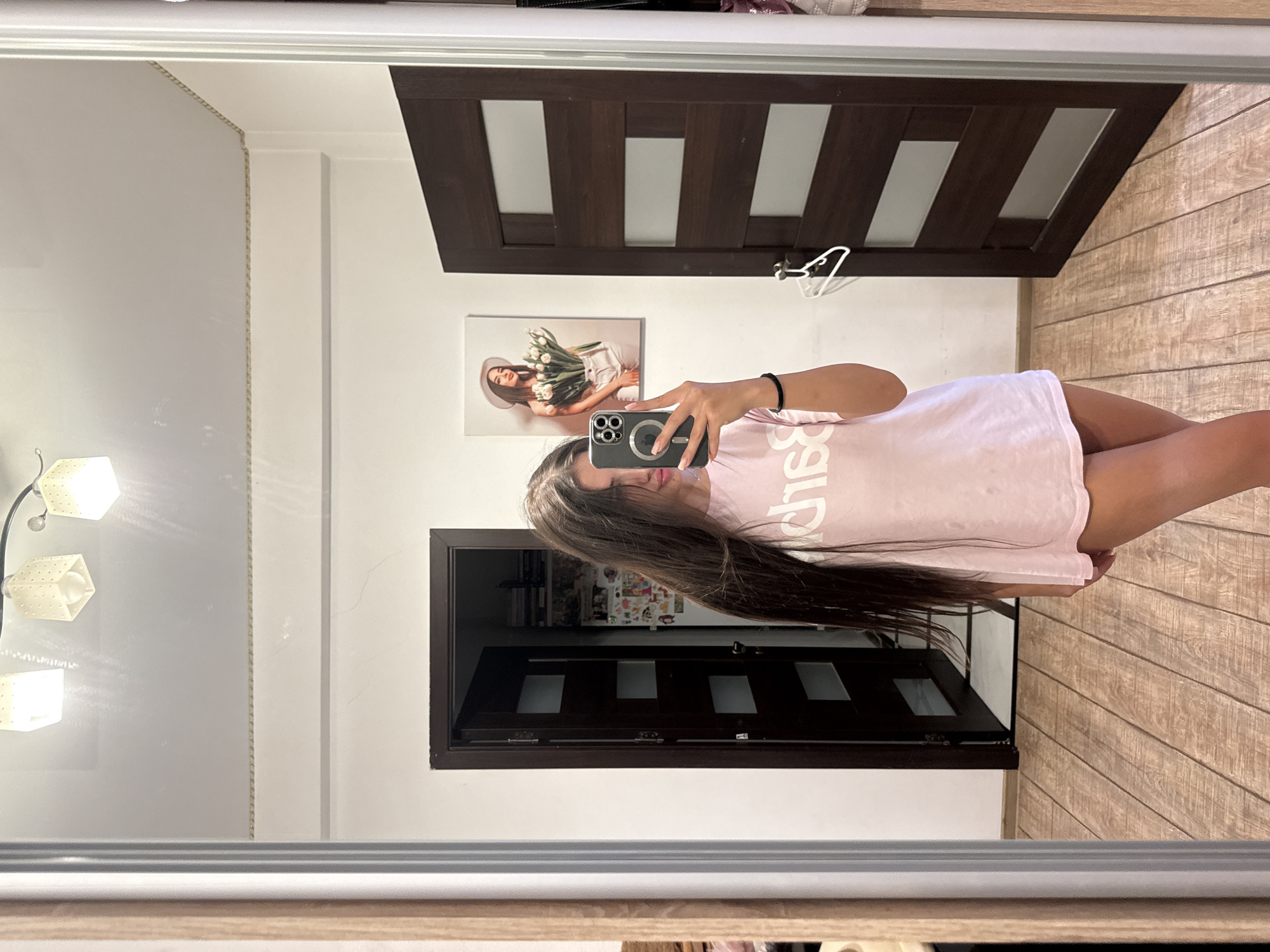 A young woman with long dark hair takes a mirror selfie in a room. She is wearing a light pink t-shirt and is standing on a wooden floor. In the background, there are two dark brown doors, one open and one closed, and a painting of a woman holding tulips on the wall. The room appears to be a bedroom or dressing area. The image does not provide any specific location details within Chișinău, Moldova.