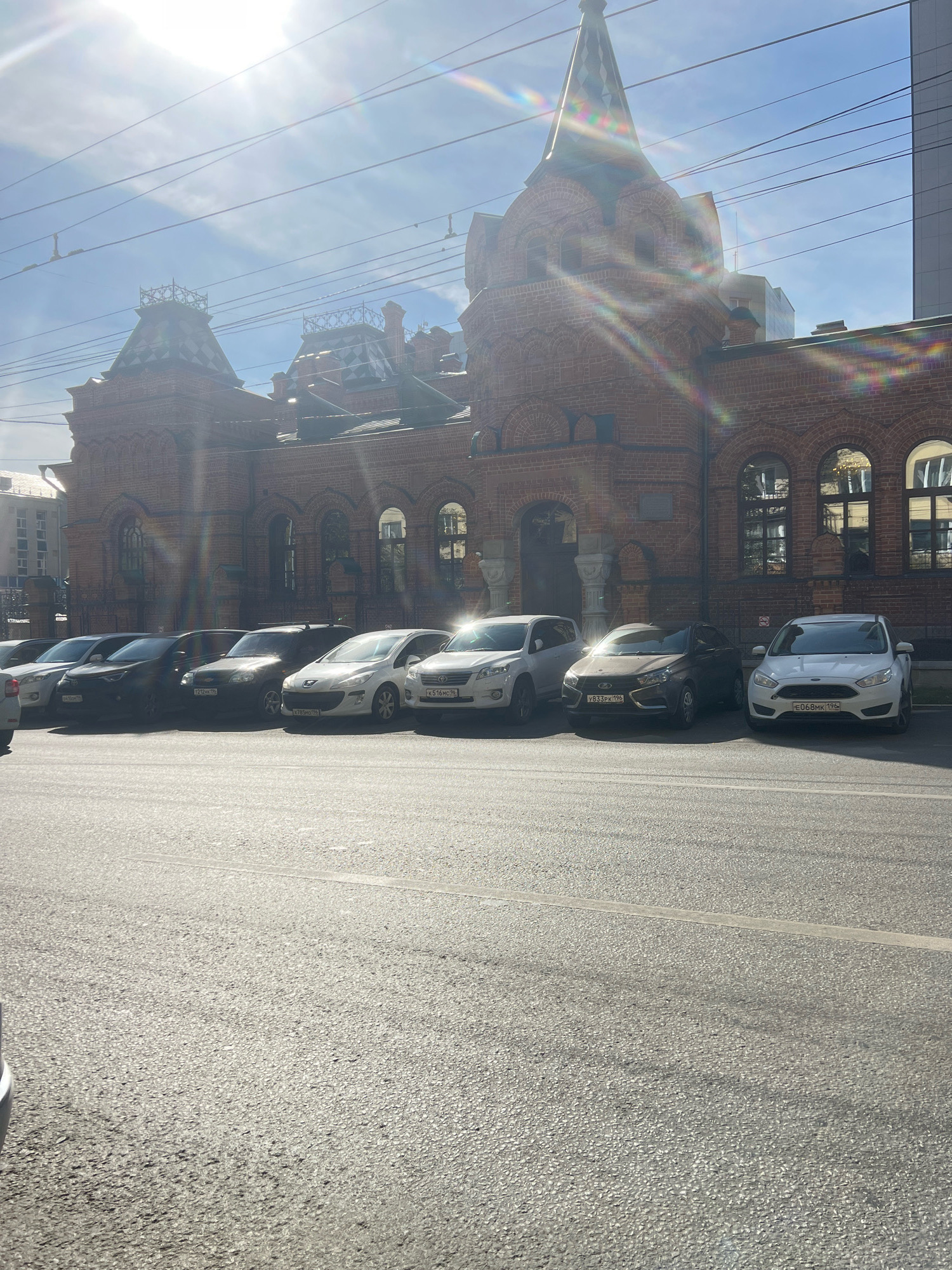 The image shows a street scene in Yekaterinburg, Russia, with a historic brick building in the background. The building features arched windows, decorative gables, and a prominent spire. In front of the building, several cars are parked along the side of the road. The sun is shining brightly, creating lens flare and casting shadows on the asphalt. Overhead, power lines are visible against the blue sky. The scene appears to be a typical urban environment with a mix of historical architecture and modern infrastructure.