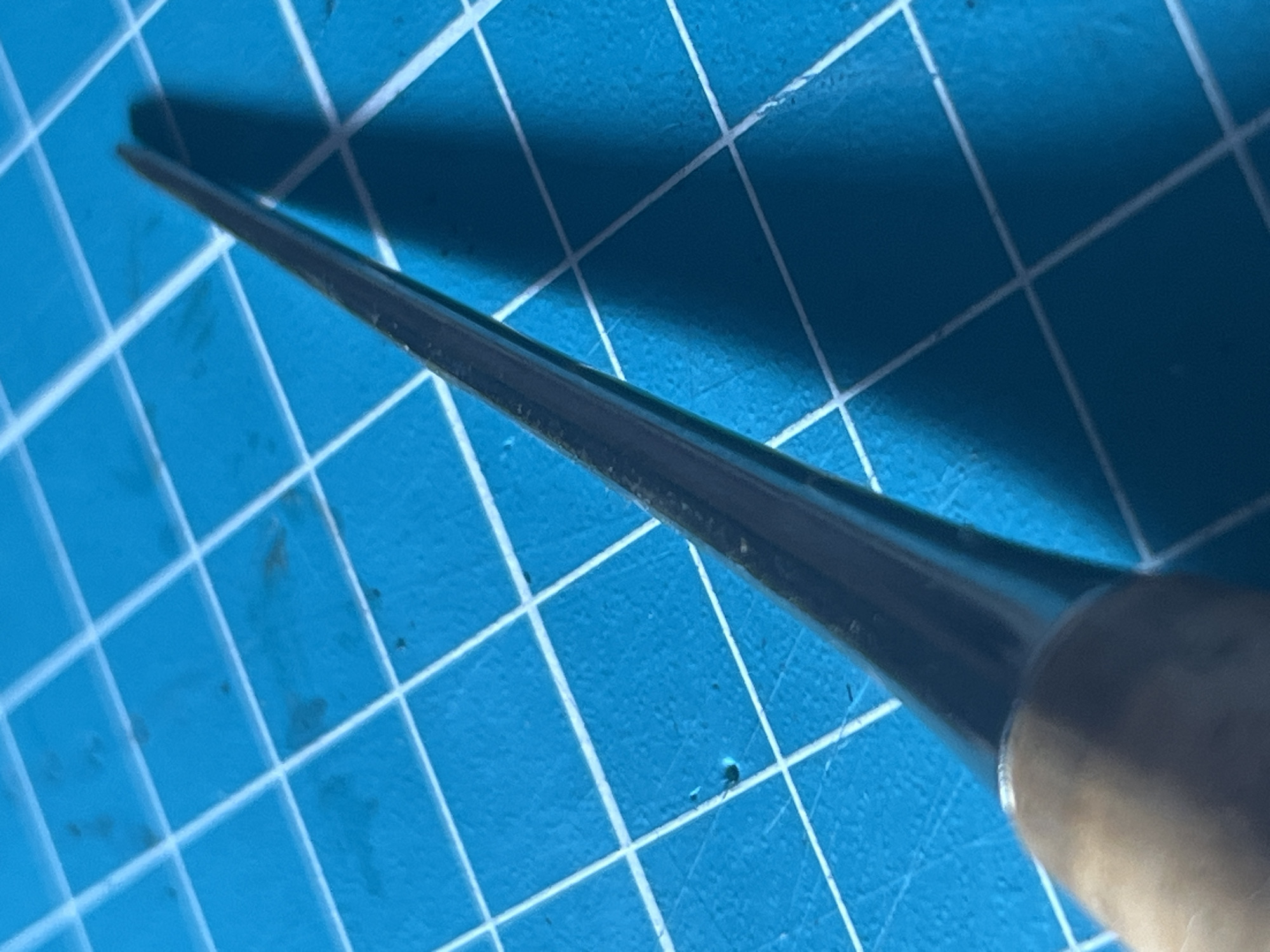 The image shows a close-up, high-angle view of a pointed tool, possibly an awl or a scribe, lying on a blue grid cutting mat. The tool has a wooden handle and a metallic, tapered point. The cutting mat has white grid lines, and some dark specks are visible on the mat. The lighting creates a shadow of the tool on the mat. There is no visible indication of the city of Novy Urengoy, Russia, or any people. The scene appears to be a still-life arrangement of a crafting or repair tool.