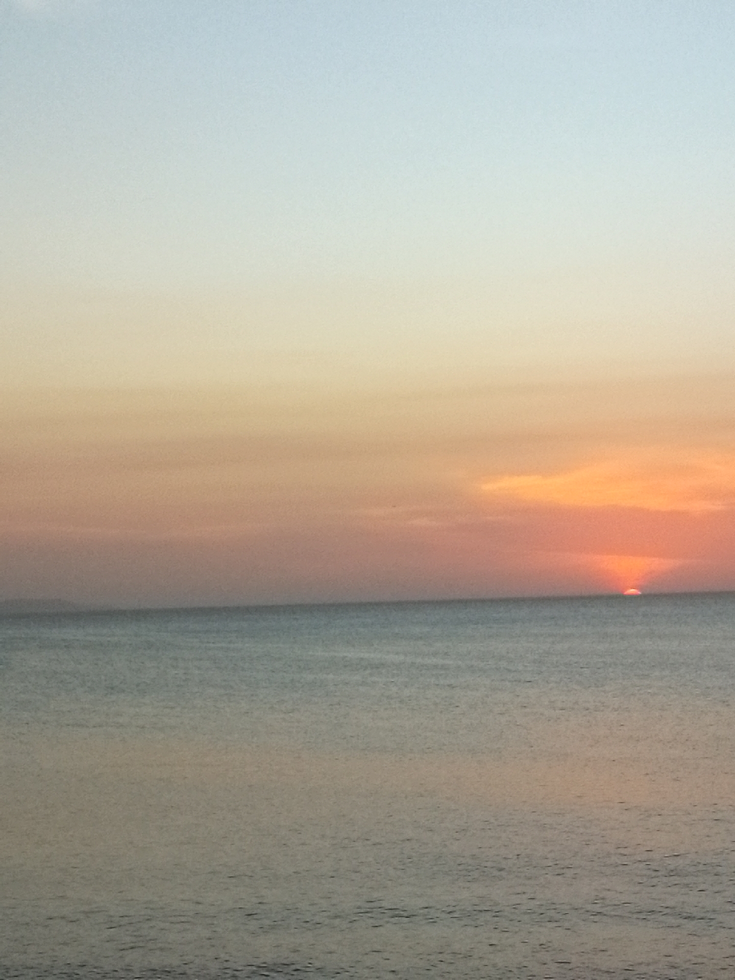 This media file depicts a serene seascape at either sunrise or sunset. The horizon line cleanly separates the sky from the ocean.
The sky exhibits a soft and expansive gradient of colors. At the top, it is a pale, clear blue, gradually transitioning downwards into muted yellows, soft oranges, and delicate pinks closer to the horizon. Towards the right side of the frame, near the horizon, the sun is visible as a bright, fiery orange-red semicircle, partially submerged below the water line. A faint band of warmer, reddish-orange clouds hovers directly above the visible portion of the sun, enhancing the glowing effect.
The ocean surface is calm with gentle, subtle ripples spread across its entirety. The water is a tranquil blue-grey, reflecting the soft hues of the sky and creating a sense of depth. A very faint, indistinct landmass or distant shoreline can be vaguely perceived on the far left side of the horizon, but the overwhelming impression is that of an open, vast sea.
The time of day is clearly dawn or dusk, characterized by the low position of the sun and the warm, blended colors of the sky. The weather appears clear and calm, with minimal cloud cover allowing for a clear view of the sun. The scene evokes a sense of tranquility and natural beauty. There are no people, animals, man-made objects, or visible text in the image. The photograph itself has a slightly grainy texture.