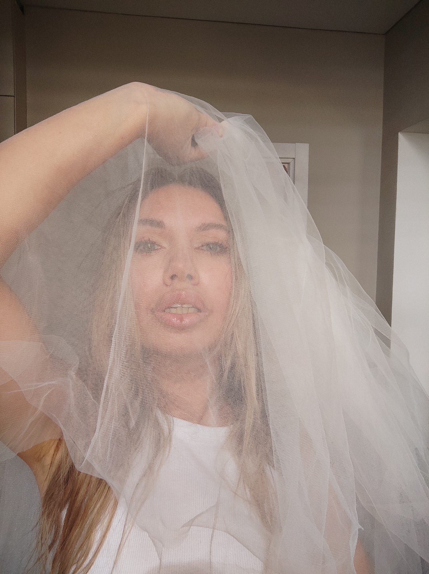 A person with light brown hair is partially visible through a white semi-transparent veil. The person is wearing a white top. The background includes a neutral-colored wall and a partial view of a white door or cabinet. The person's right arm is raised, holding the veil overhead. It is impossible to ascertain the location as Votkinsk, Russia, based on the image alone.