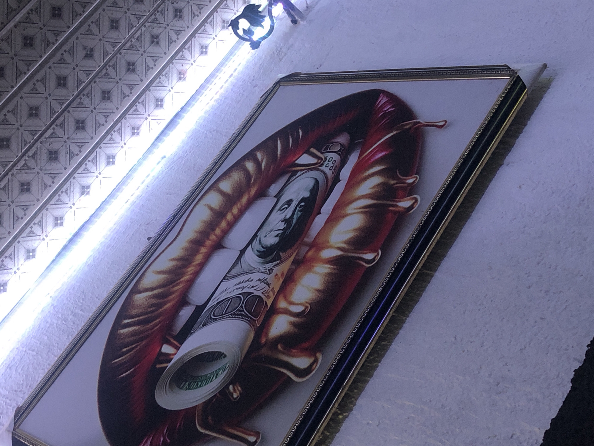 A framed artwork featuring a close-up depiction of red lips with a roll of U.S. dollar bills held within them is mounted on a textured, white wall. The artwork is framed with a multi-layered border, including gold and dark blue sections, and is slightly tilted. Above the artwork, a bright LED strip light illuminates the scene. The background wall has a textured pattern, and a decorative metal piece, shaped like a stylized leaf or creature, is visible on the upper right side. The overall impression is of a stylized and somewhat abstract artistic representation of wealth and desire.
