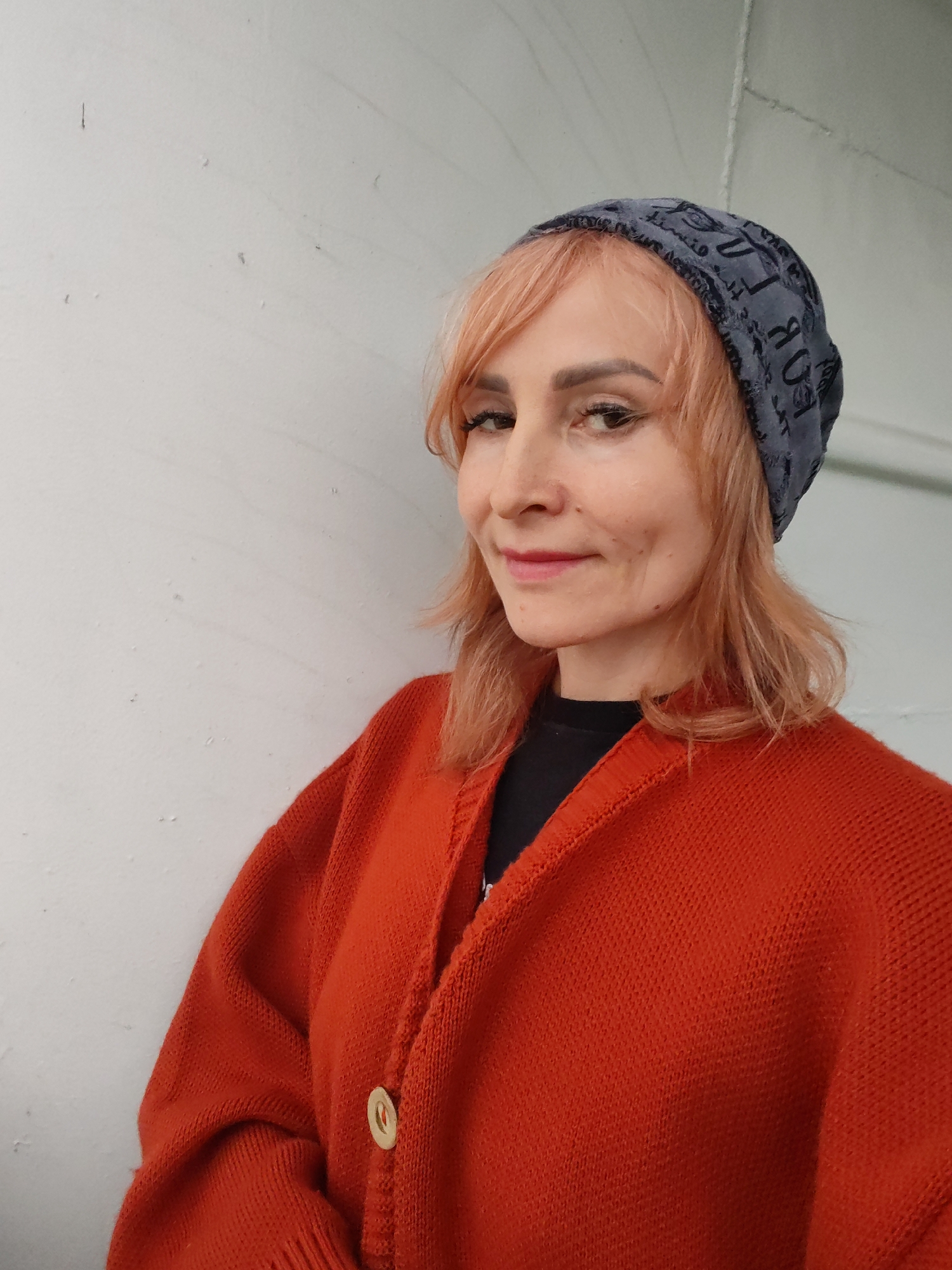 A medium shot shows a woman with shoulder-length, peachy-blonde hair, wearing a grey beanie with a black, patterned design. She is also wearing a bright orange cardigan over a black shirt. The cardigan has a single, light-colored button. The woman is leaning against a textured white wall, which appears to be indoors or in a sheltered outdoor space. Her expression is a soft smile, and her eyes are looking towards the viewer. The lighting suggests it is daytime. The setting appears to be a simple, unadorned environment. The pattern on the beanie contains text, but it is not legible in the image.
