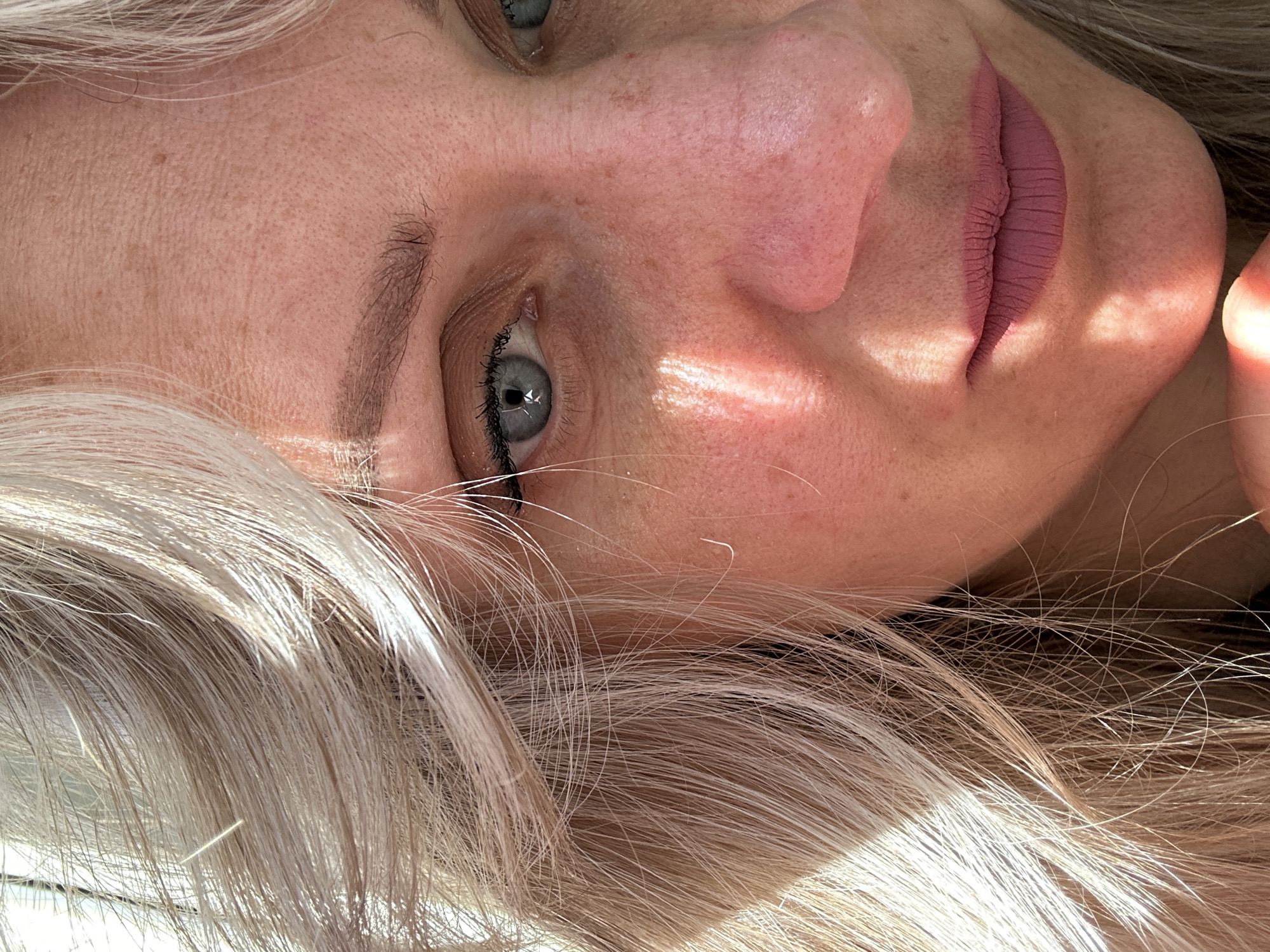 A close-up, rotated portrait captures the upper half of a woman's face. The image is oriented with the woman's head tilted about 90 degrees clockwise, so her right eye is in the lower center and her mouth is towards the right side of the frame. She has fair skin with visible pores, fine lines, and scattered freckles. Her left eye (the one more prominently visible) is open, revealing a light blue or gray iris, outlined with dark eyeliner and mascara. Her eyebrow on that side is well-defined and dark.
Strong, directional sunlight casts prominent highlights and shadows across her face. A bright highlight illuminates her nose bridge and the area above her eye, while another band of light strikes her chin and lips. Her lips are full and adorned with a matte, dusty rose or mauve lipstick, showing natural creasing.
Her hair, which appears to be platinum blonde or very light gray, frames the lower and upper left portions of the image, catching the sunlight and appearing luminous. Individual strands of hair are clearly visible. The overall impression is a detailed, intimate, and sunlit portrait focusing on facial features and skin texture. No specific activity beyond posing for the photograph is discernible. There is no visible text. The location context is Venafro, Italy.