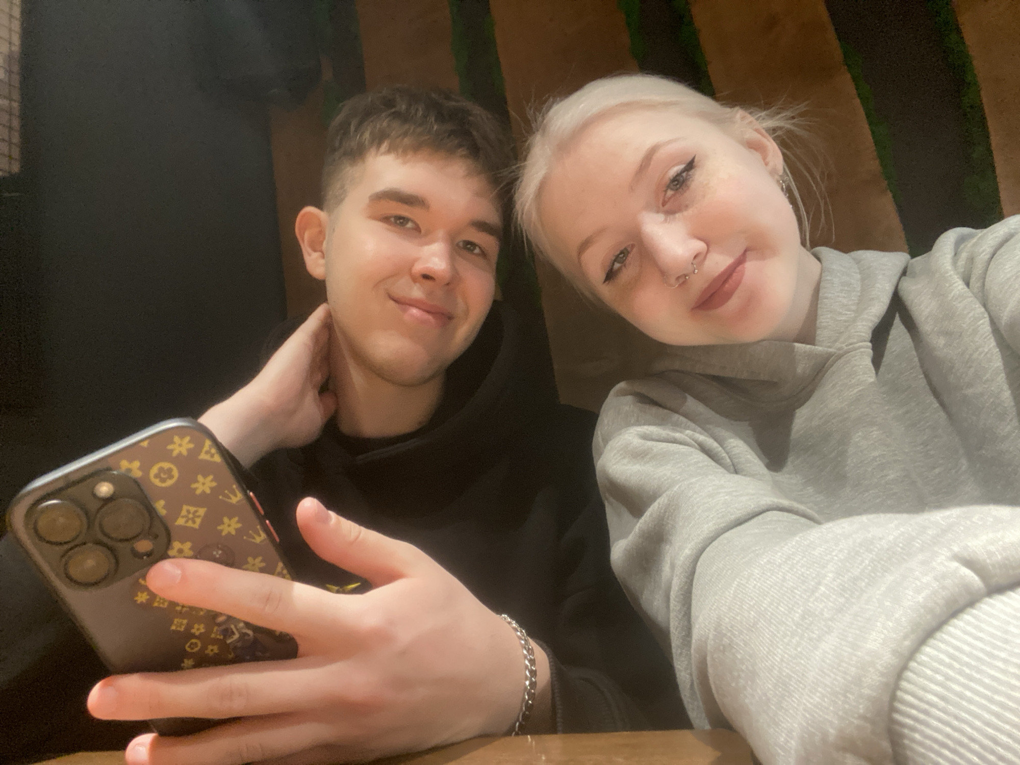 A selfie of two young adults, a male and a female, seated indoors at a table. The male is on the left wearing a black hoodie and holding a smartphone with a brown patterned case. The female is on the right, wearing a grey hoodie, earrings, and nose rings. They both smile at the camera. The background is out of focus but appears to be an interior space with dark walls and possibly some vertical green accents. The location is purported to be in Izhevsk, Russia.