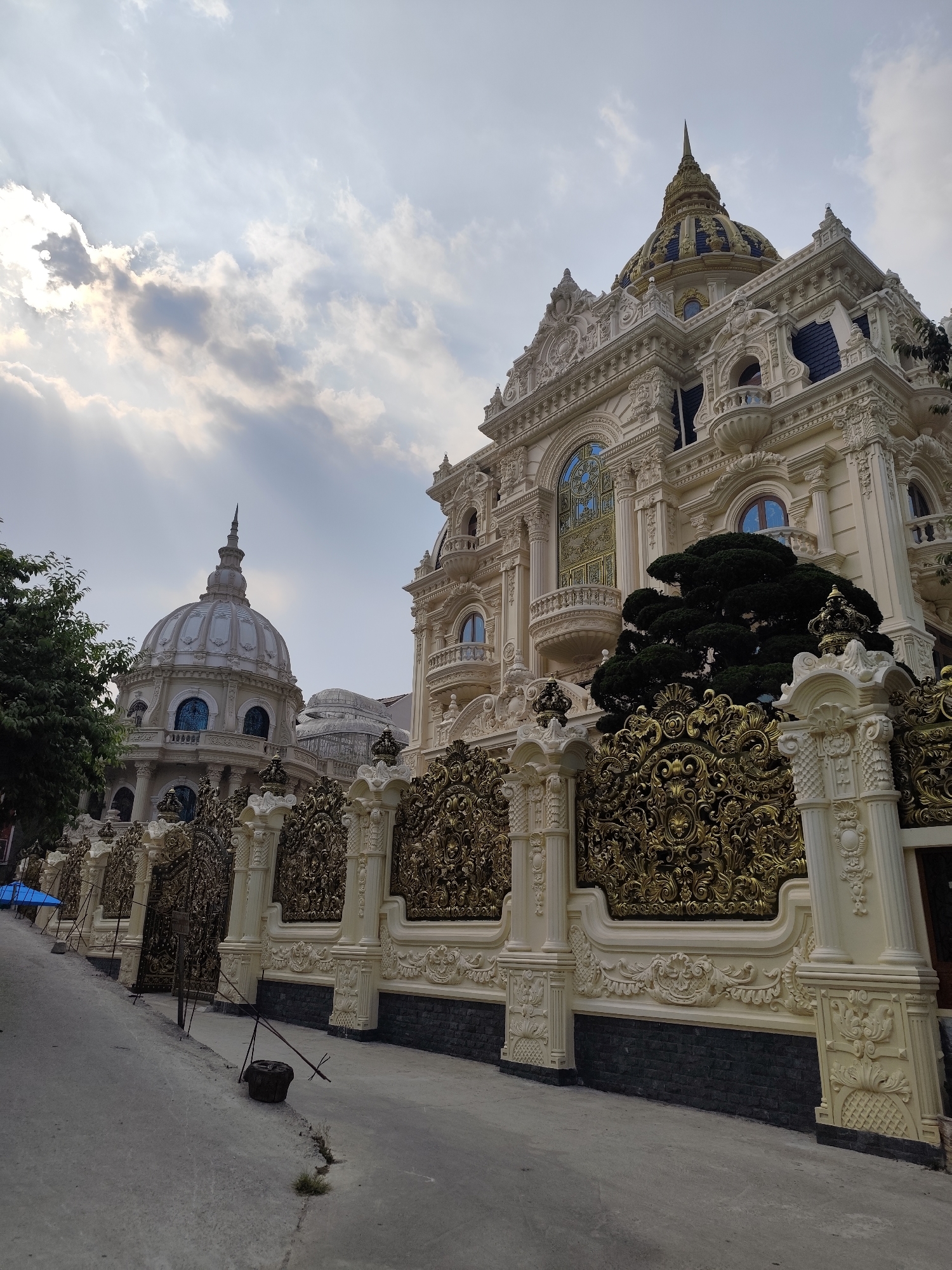 The image showcases a lavishly ornate building with domes, located in Da Lat, Vietnam. The building is characterized by intricate architectural details, including sculpted columns, decorative cornices, and gilded fence work. A large, central structure features a blue and gold dome, while a smaller dome-topped building is visible to the left. The fence is made of ivory and gold columns and filigree. The sky above is cloudy, with diffused sunlight. A narrow concrete sidewalk runs along the base of the fence. The environment suggests an urban or suburban setting in Da Lat, Vietnam. There are no visible people in the image.