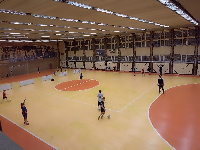 An elevated view captures an indoor sports gymnasium in Fedorovka, Russia. The facility features a light orange and orange two-toned synthetic court with white demarcation lines, including a central oval and a large curved boundary on the right. The high ceiling is fitted with numerous horizontal fluorescent light fixtures. The upper walls consist of a series of windows, while the lower sections are paneled in beige with a dark patterned trim. A large mural depicting athletes is visible on the far left wall, beneath which a dark sports goal is positioned. A digital scoreboard displaying "0:04" and other data is mounted on the far wall. Approximately 20 individuals, predominantly young males, are present on the court. Most are dressed in athletic attire (t-shirts, shorts) and are engaged in activities consistent with soccer or futsal training, with several handling soccer balls. Multiple white, low barriers or small goals are deployed in the mid-ground. An adult male in dark athletic wear stands on the right side of the orange court area, observing the activity.