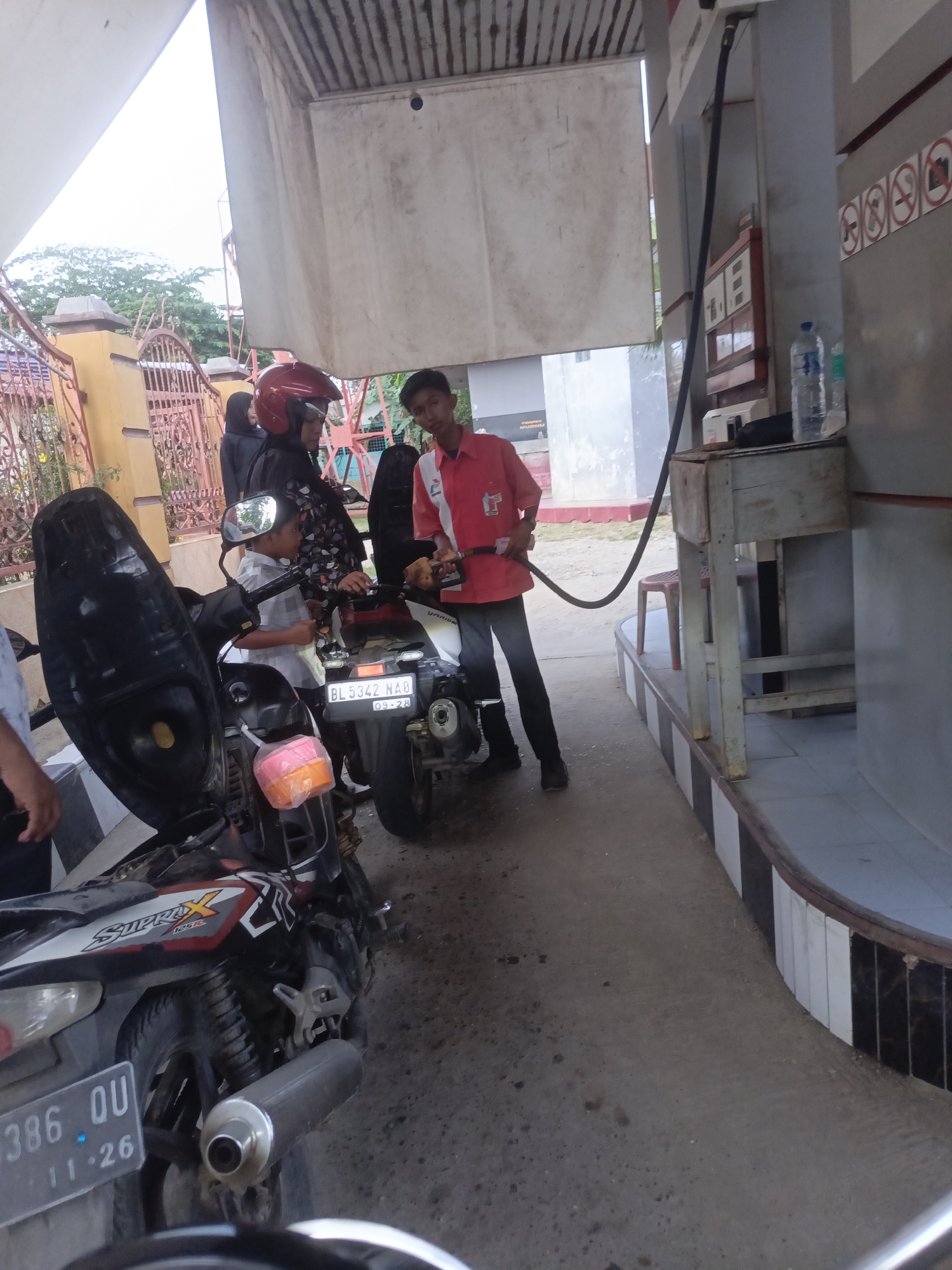 The image depicts a scene at a gas station in Lhokseumawe, Indonesia. A young boy, wearing a grey checkered shirt, is standing next to a motorbike, appearing to be involved in fueling it. A gas station attendant, dressed in a red uniform with a logo, is holding a gas nozzle and appears to be dispensing fuel into the motorbike. Two women are also present. One woman is wearing a red helmet and a patterned top, and the other, mostly obscured, is wearing a dark headscarf. The environment includes a gas pump with a hose, a covered awning, and the street or pavement of the gas station. The motorbike in the foreground has a license plate that reads "386 QU 11-26" and the brand name "Supra X 125 FI" is visible. The background shows fencing, trees, and buildings, consistent with an urban or semi-urban setting in Indonesia. The lighting suggests it is daytime, possibly overcast.