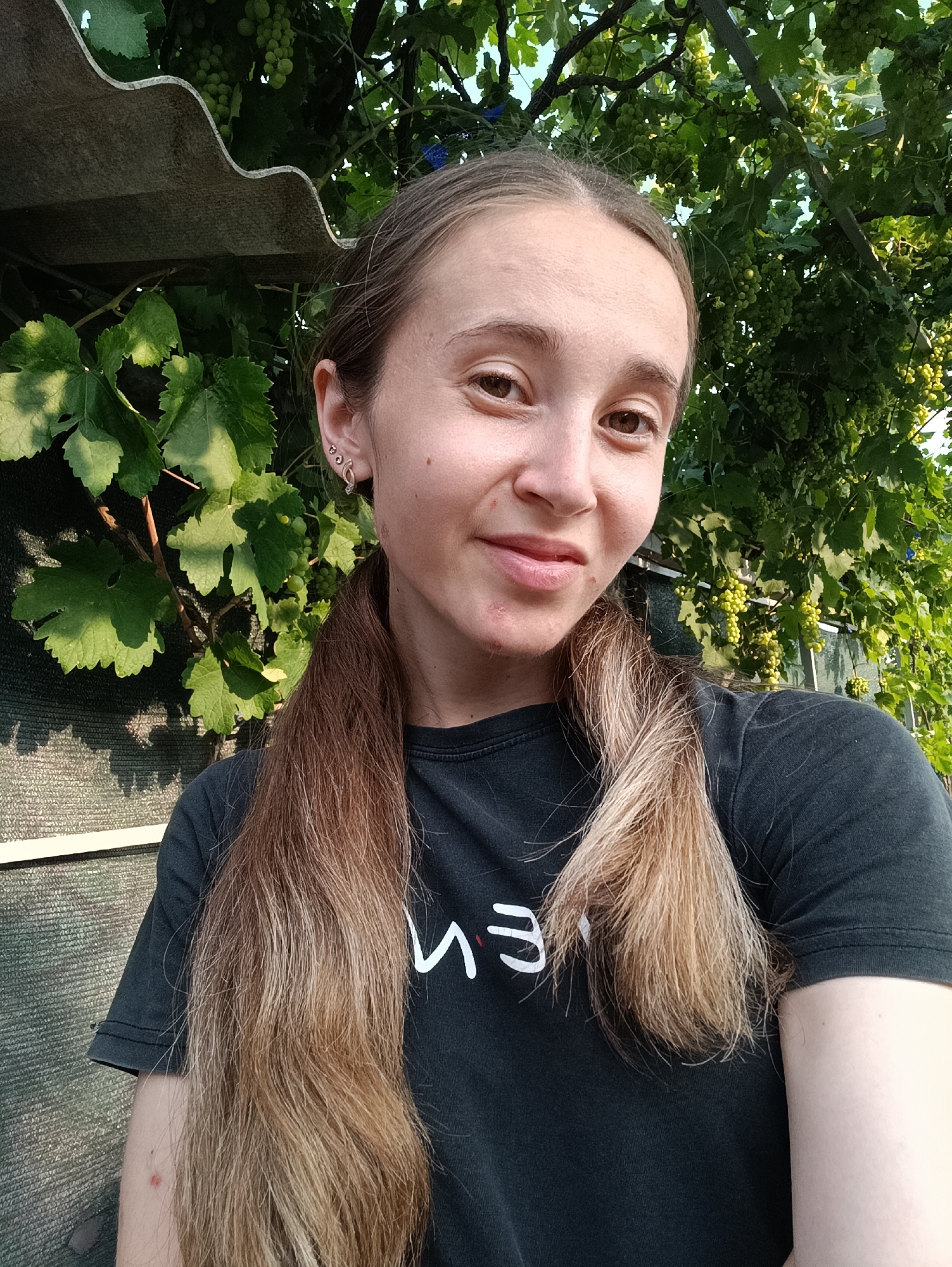 The image is a selfie taken outdoors during the day, with a young woman with long, braided blonde hair in the foreground. She is wearing a dark t-shirt with white lettering. Behind her, there are green grapevines with clusters of unripe green grapes. To the left, a corrugated roof panel is visible, and behind that, a woven fence. The setting appears to be a residential area or garden. The image does not provide specific geographic information that would place it in Vypasne, Ukraine.