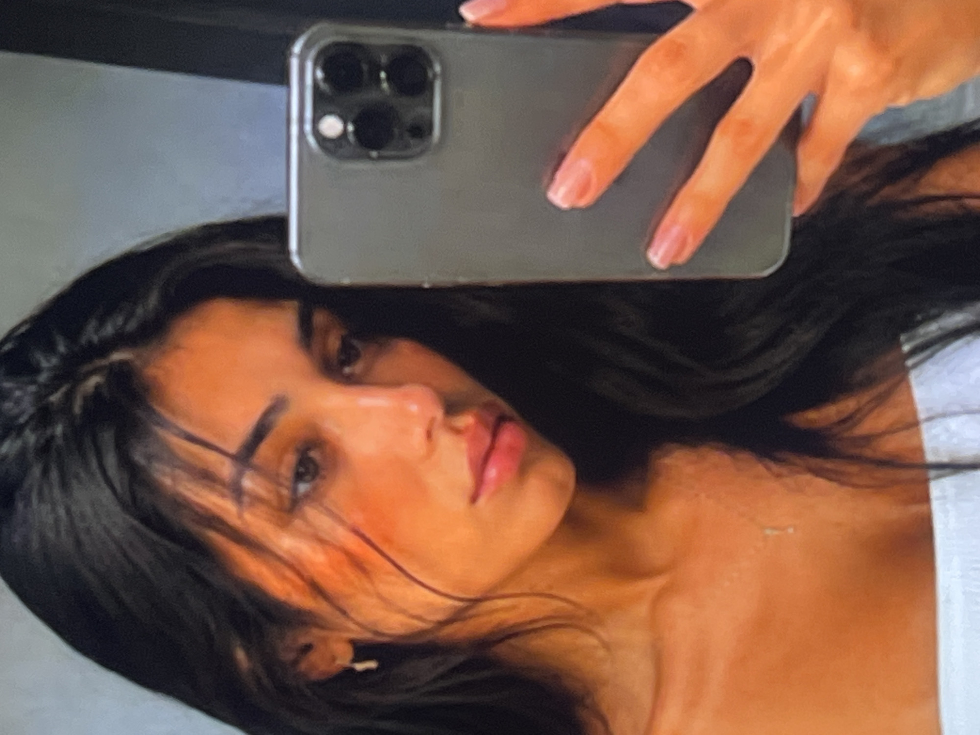 The image shows a close-up, low-angle selfie taken by a person holding a smartphone. The subject is a young person with dark hair and fair skin, wearing a white top. They are looking at the camera with a neutral expression. The smartphone, a modern model with a prominent camera module, is visible in the upper part of the frame, held by a hand with manicured nails. The background appears to be a plain, light-colored wall. The lighting suggests an indoor setting, possibly a bathroom or bedroom, and the time of day is indeterminate. The overall tone of the image is casual and personal, typical of a selfie.