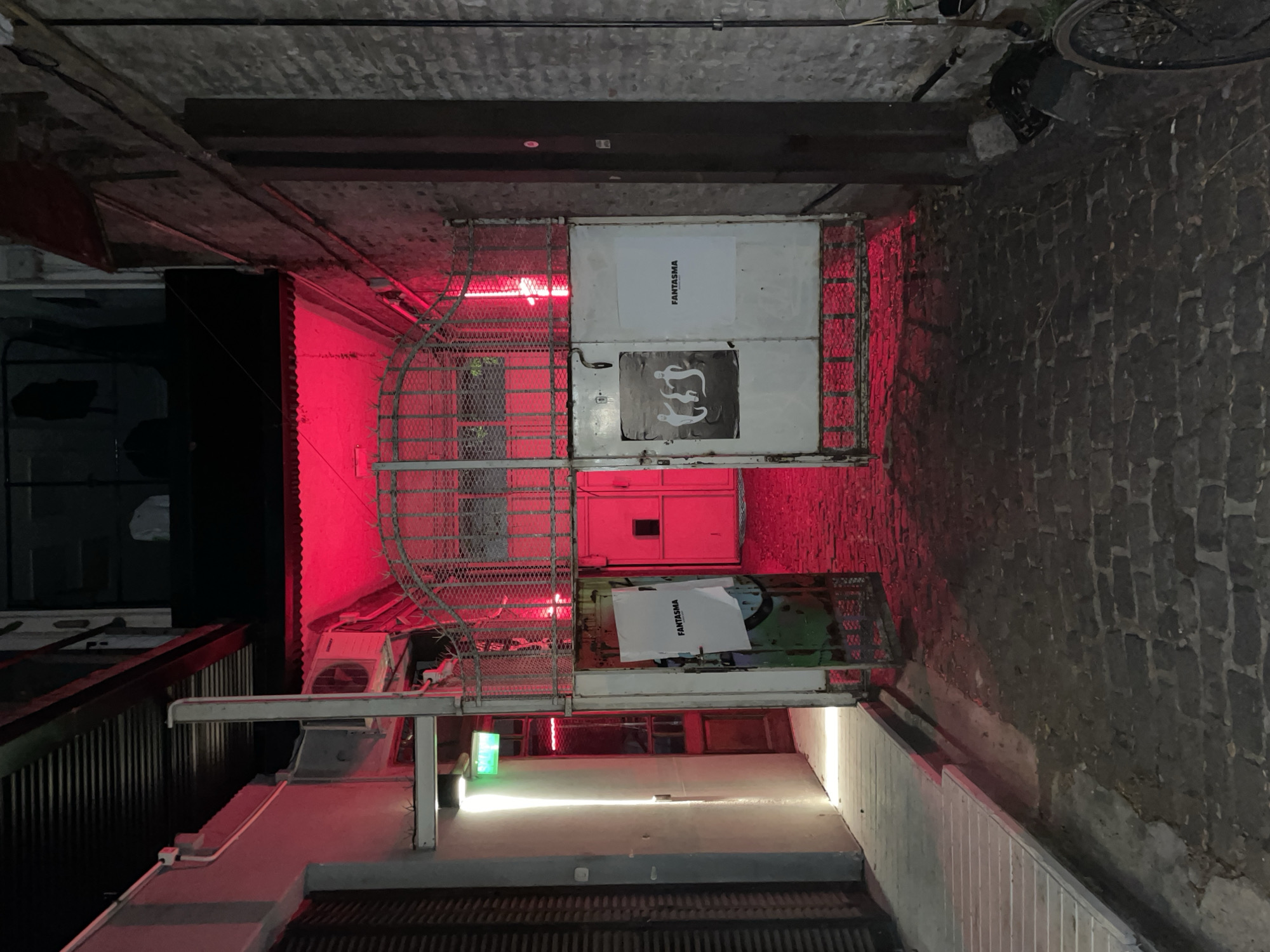 The image depicts a narrow urban alleyway at night, illuminated by a striking combination of red and white artificial lights. The setting suggests an industrial or hidden city space.
On the left side, a strong red light emanates from above, casting a vibrant crimson hue across the wall, which appears to be constructed of vertical metal slats in the foreground and a smoother surface further back. An outdoor air conditioning unit is visible on a ledge on this left wall. Deeper into the alley on the left, a soft green light also provides illumination.
The right side of the alley features a rough, textured brick wall extending into the distance. A bicycle is parked against this wall in the upper right background. The overhead ceiling is dark and textured, with visible pipes and beams running across it. The ground is paved, displaying varying textures that could be concrete and cobblestone.
A prominent metal gate or cage-like structure spans the width of the alley in the mid-ground, acting as a barrier. This structure incorporates metal mesh panels on its sides and solid white panels in its center. Two rectangular signs are affixed to these central white panels. Both signs feature the word "FANTASMA" in black, sans-serif text; one is oriented vertically, and the other horizontally. Below the horizontal "FANTASMA," a dark poster or sticker displays the white outlines of two stylized, monkey-like figures.
There is no human activity present, contributing to a quiet, somewhat dramatic atmosphere. The scene is clearly set at night, with the intense red lighting creating an edgy or mysterious mood, possibly indicating the entrance to a nightlife venue.