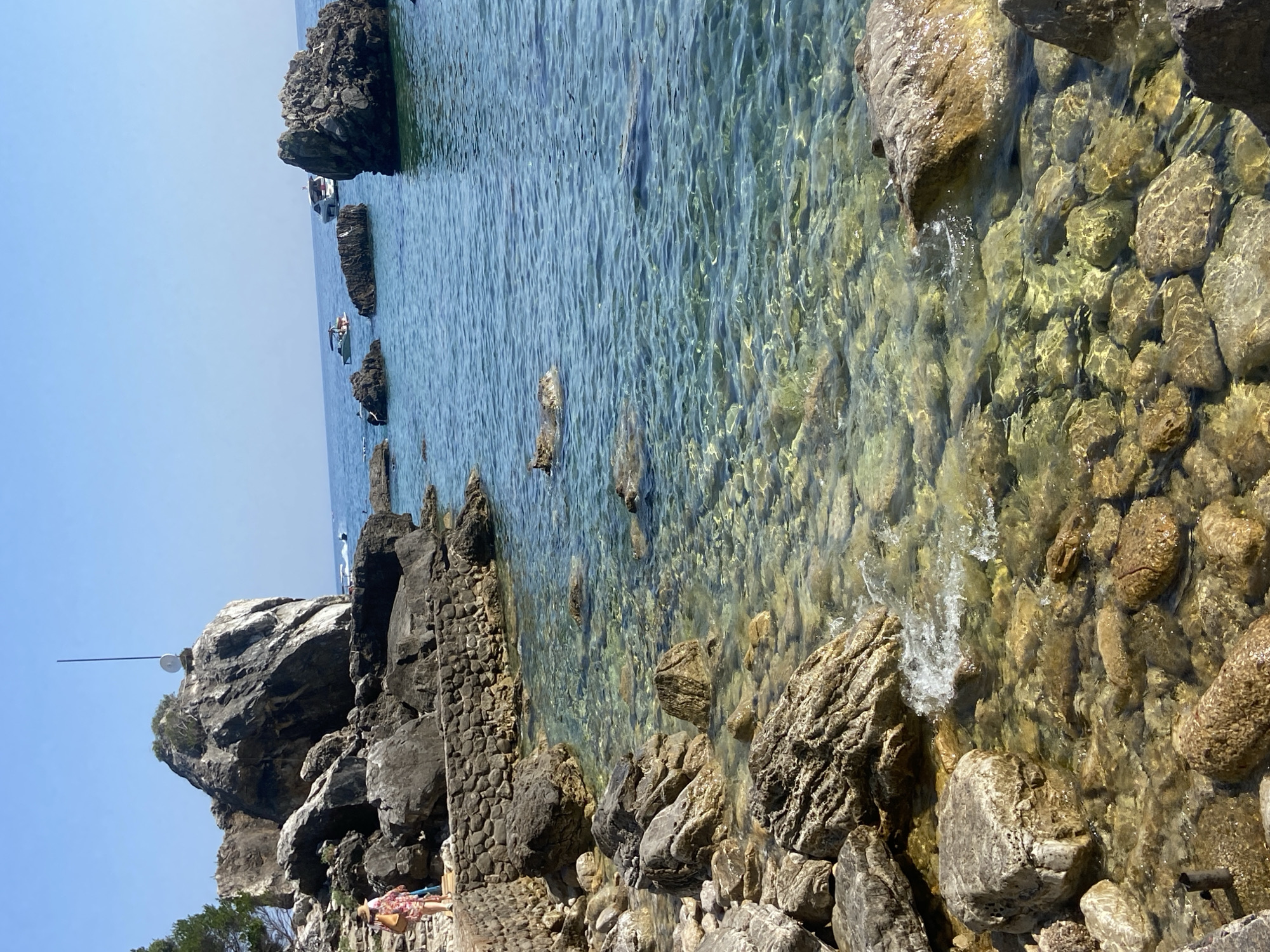 A person walks along a stone pathway next to a rocky coastline under a clear blue sky. The pathway is constructed of rough stones and leads towards a large boulder. Beyond the pathway and rocks is the bright blue water of the sea, which is clear enough to see the rocky bottom. Several boats are visible on the water in the distance. The overall environment is a scenic coastal landscape.
