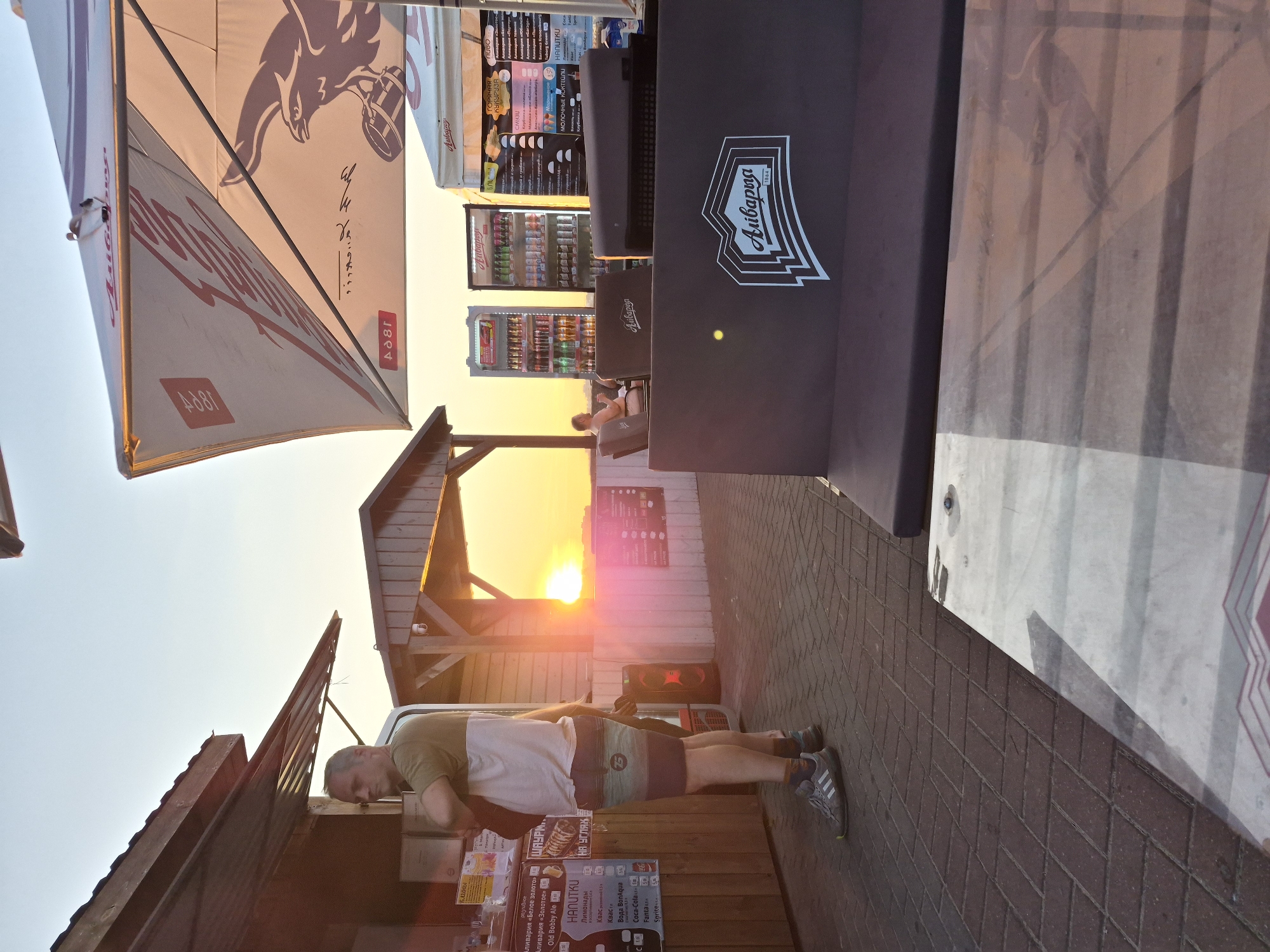 The image captures a sunset scene at an outdoor establishment, likely a bar or cafe, in Minsk, Belarus. The warm glow of the setting sun illuminates the sky, casting long shadows and highlighting the textures of the paved ground and wooden structures.
In the foreground, a man stands near a food stall, his back partially to the camera. He is wearing a t-shirt with a light-colored top and striped shorts, and athletic shoes. He appears to be looking at the menu board. To his left, a large, dark speaker is visible.
Behind the man and the stall, a rustic wooden structure with a sloped roof is visible, possibly a shelter or part of the establishment's architecture. Further back, two large glass-fronted refrigerators are filled with bottled and canned beverages. The refrigerators are branded with the "Aivaryya" logo. To the right of the refrigerators, a white tent with the "Aivaryya" logo is set up, likely another seating or serving area.
A large umbrella, also branded with "Aivaryya" and featuring an eagle graphic, is prominently displayed in the upper right portion of the image, providing shade. The umbrella's design suggests it is part of the establishment's branding.
In the mid-ground, a person is seated on a low, dark-colored sofa or bench, facing away from the camera and towards the sunset. The bench is also branded with the "Aivaryya" logo.
The overall impression is one of a relaxed, evening gathering, possibly at a venue catering to tourists or locals enjoying the outdoor atmosphere. The presence of the "Aivaryya" branding throughout the scene indicates it is a specific establishment or chain.