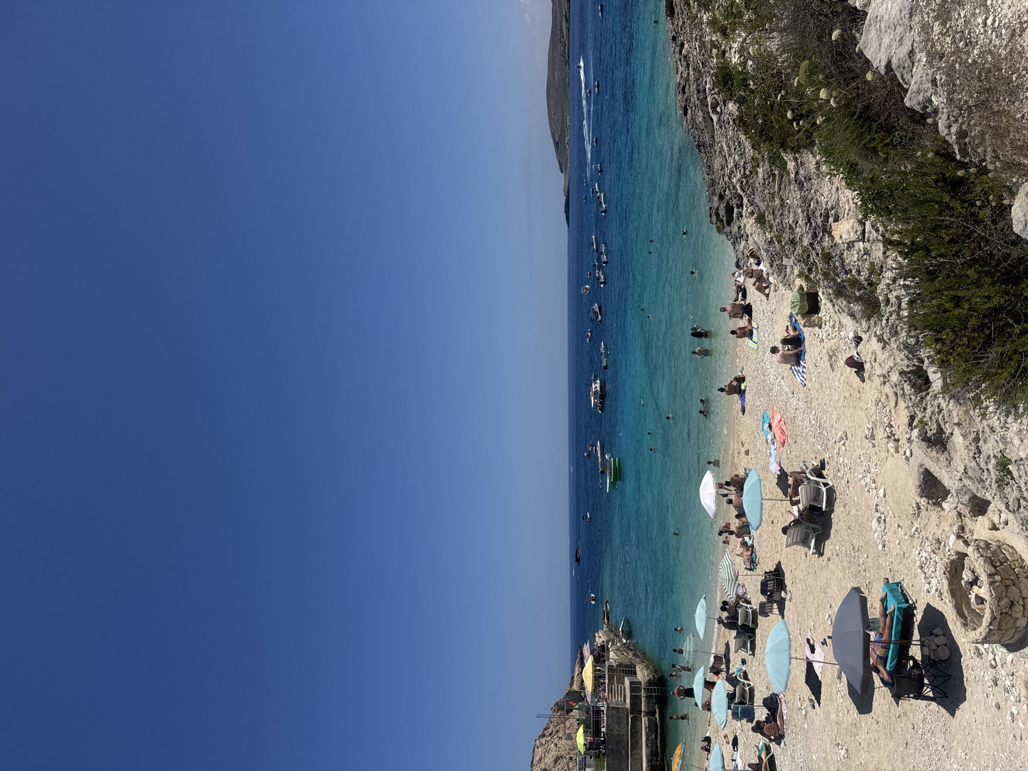 This is a high-angle, full shot of a busy beach on a clear, sunny day. The sky is a solid, vibrant blue. The sea is a bright turquoise, with many boats scattered across its surface, some appearing to be moving. The beach is a mix of sand and pebbles, with numerous beachgoers sunbathing, swimming, and relaxing under umbrellas. There are lounge chairs and towels visible on the beach. To the left, there is a rocky shoreline with some buildings and a pier. The water is clear enough to see people swimming and the seabed in some areas. The overall scene depicts a popular tourist destination in Qala, Malta.